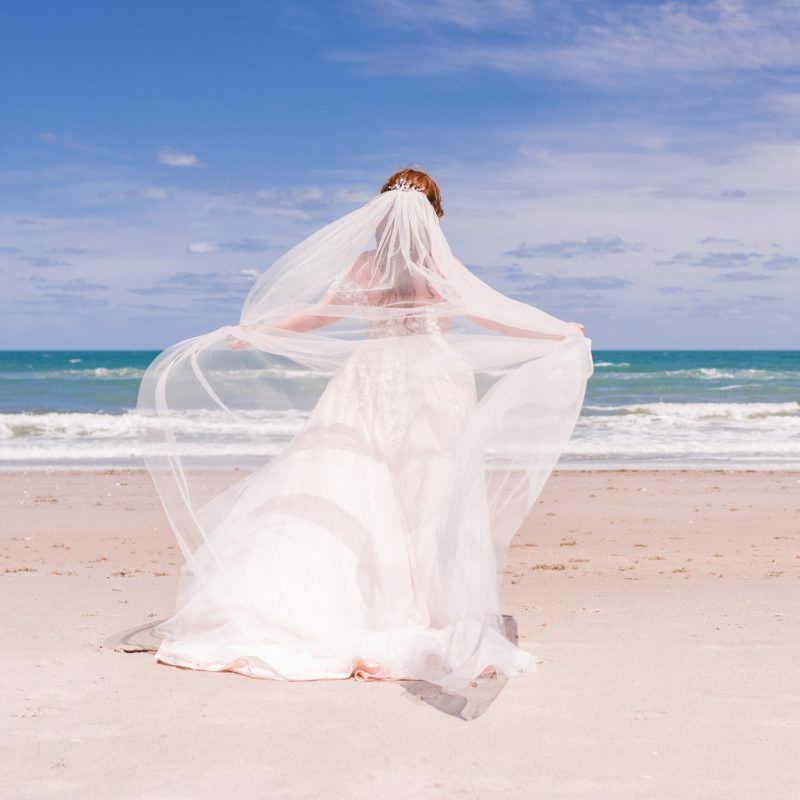 Bride facing the ocean with veil blowing in the wind on a sunny day on Melbourne Beach