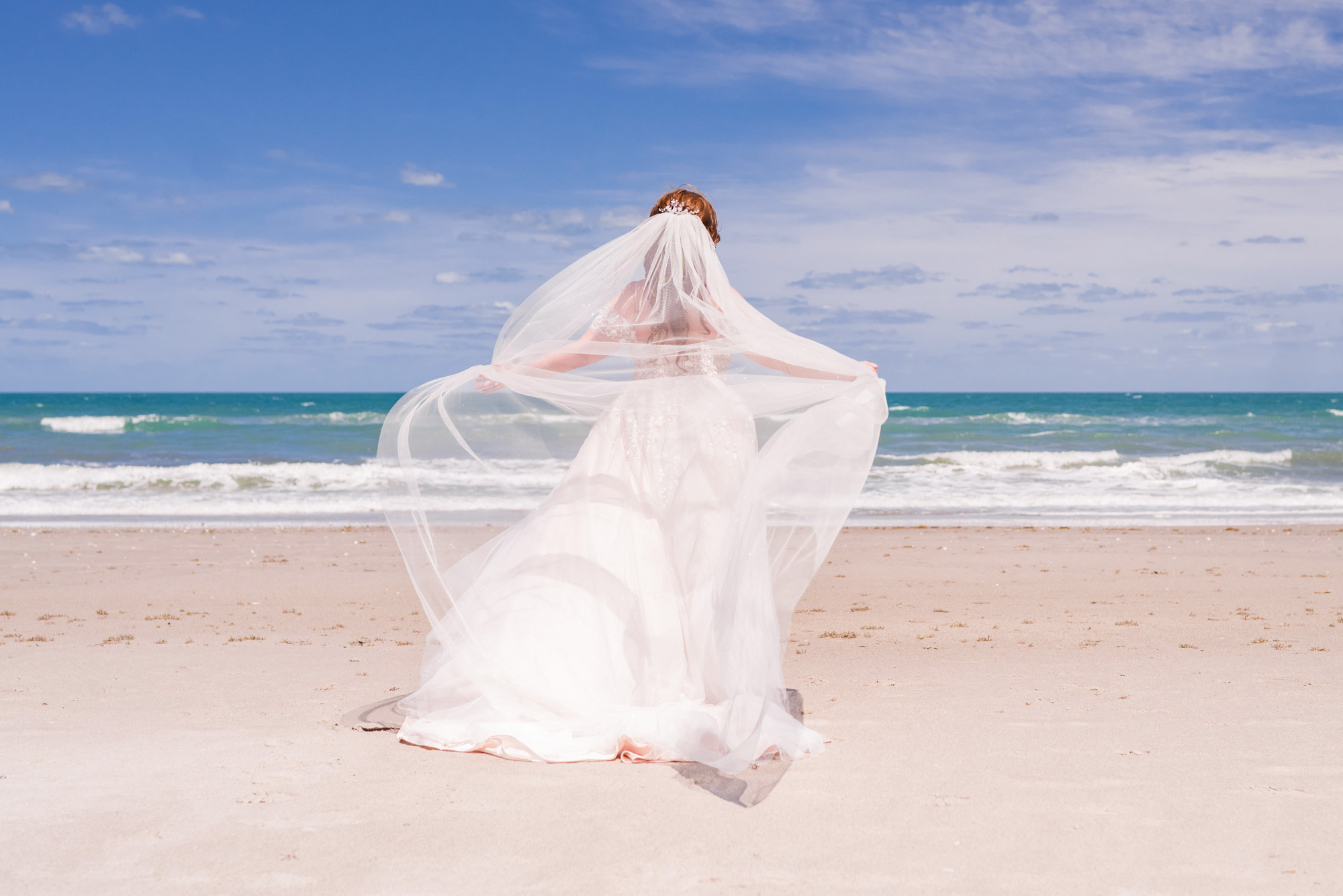 Bride facing the ocean with veil blowing in the wind on a sunny day on Melbourne Beach