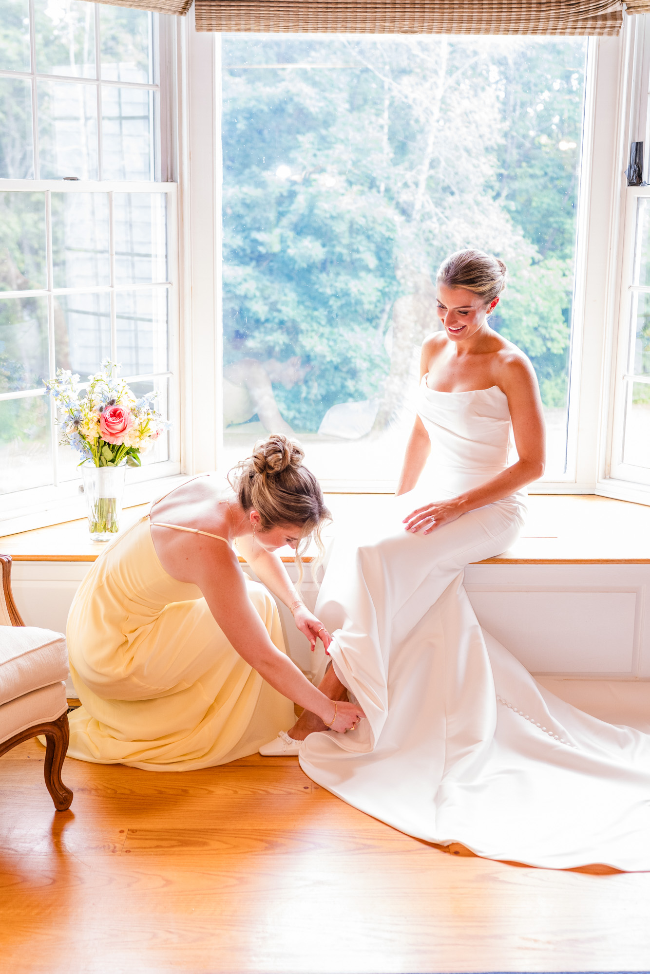 Bridesmaid helping the bride with her shoes beside a sunlit window