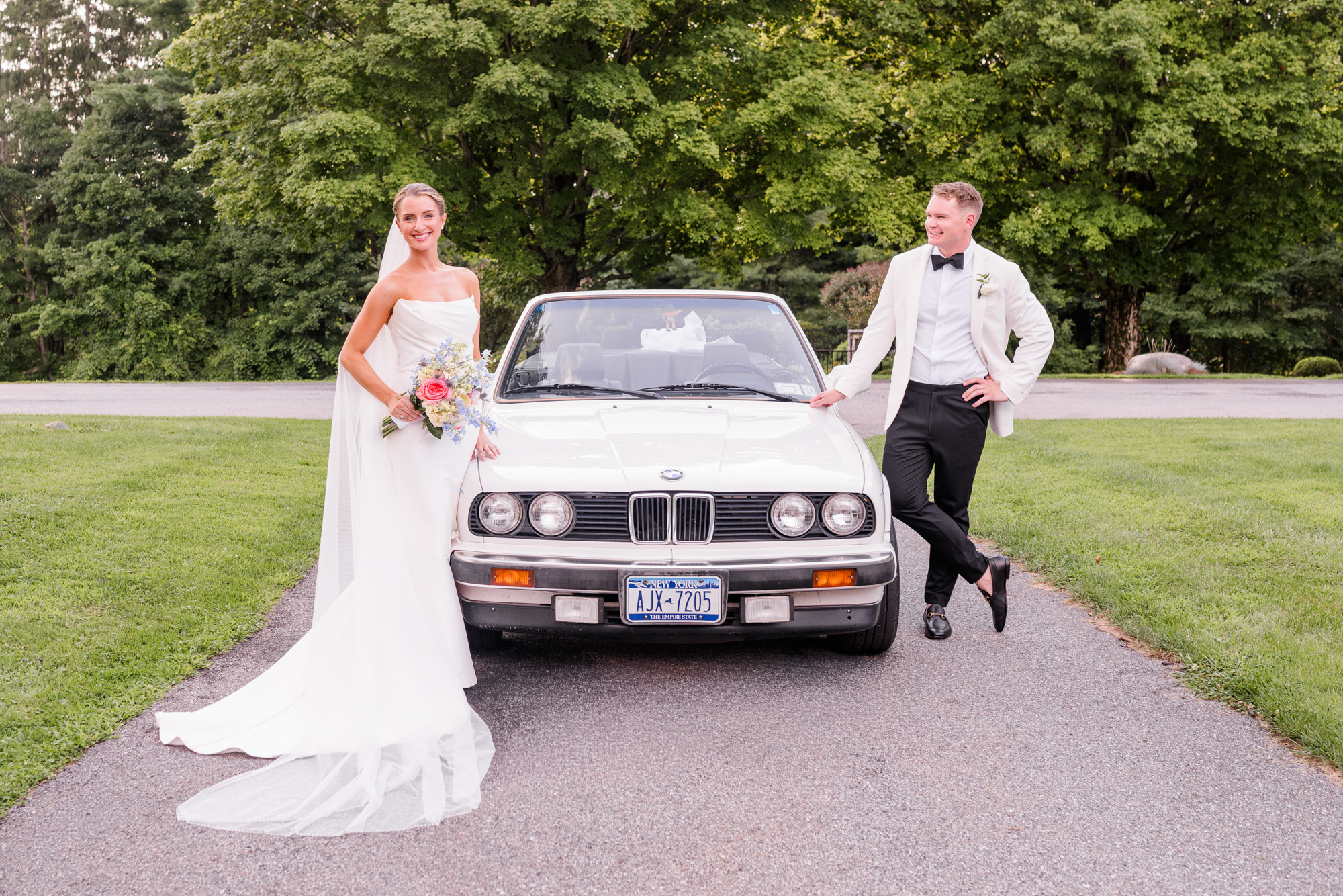 Newlyweds standing beside a vintage BMW surrounded by summer greenery