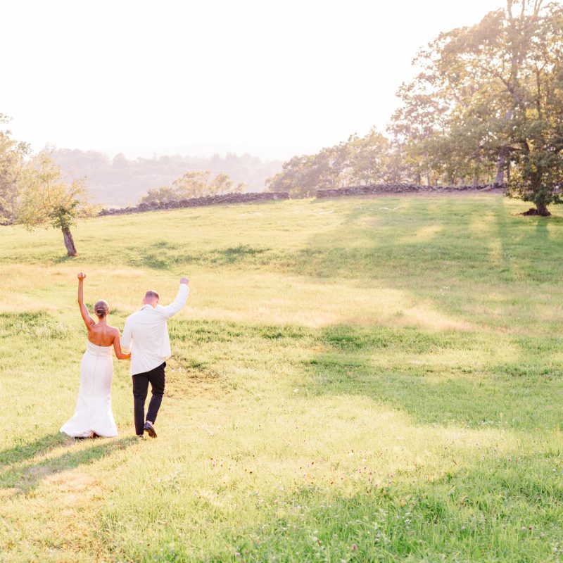 Bride and groom walking together through a glowing summer field in Pawling NYinto the sunset with their arms in the air