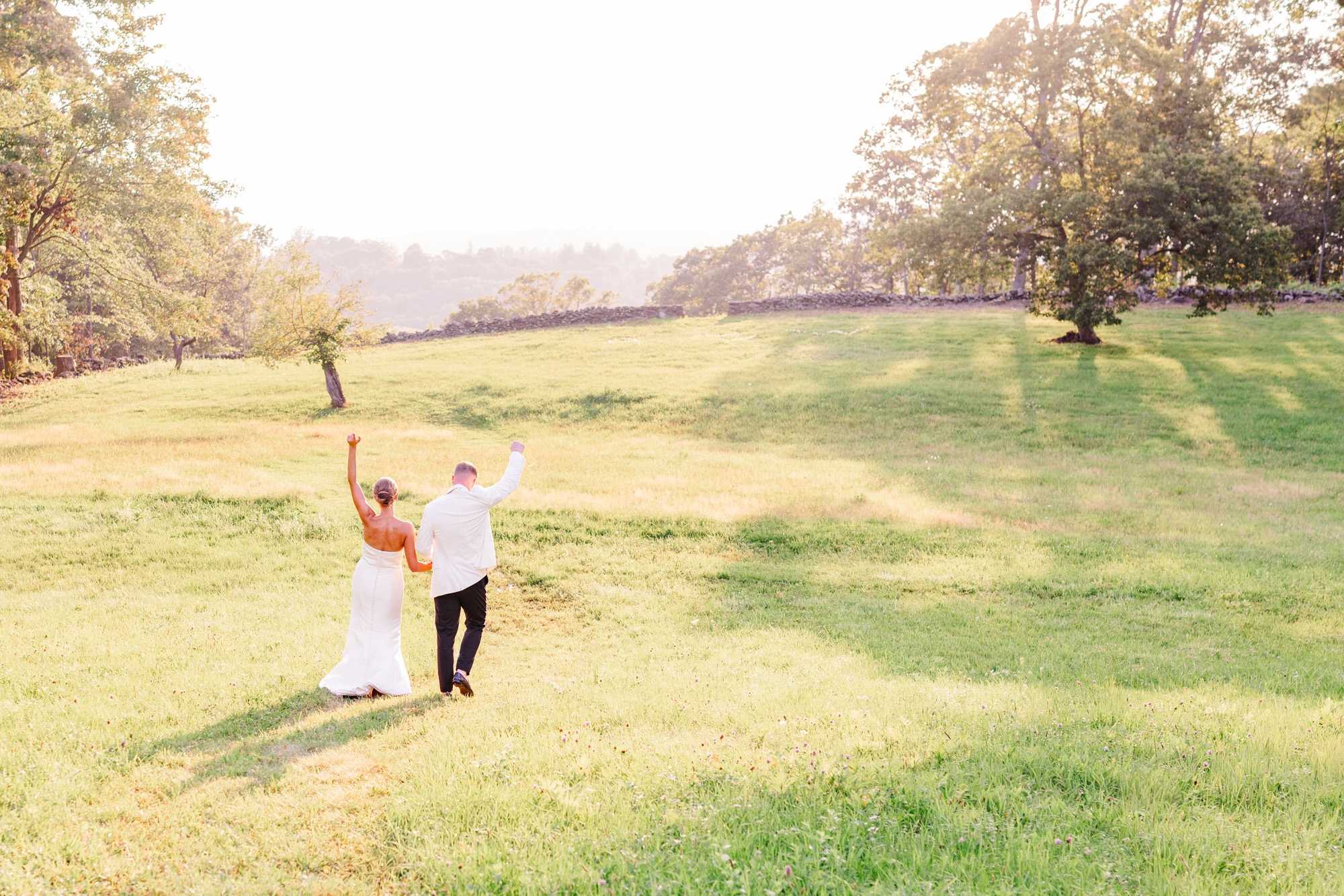 Bride and groom walking together through a glowing summer field in Pawling NYinto the sunset with their arms in the air