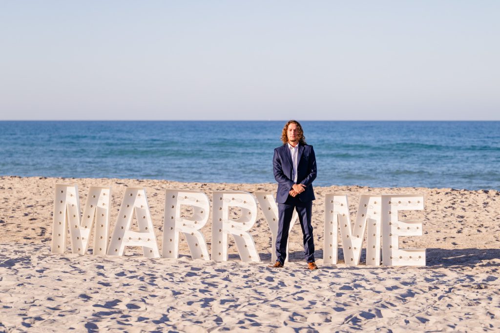 Man standing in front of giant marquee letters on the beach that say marry me