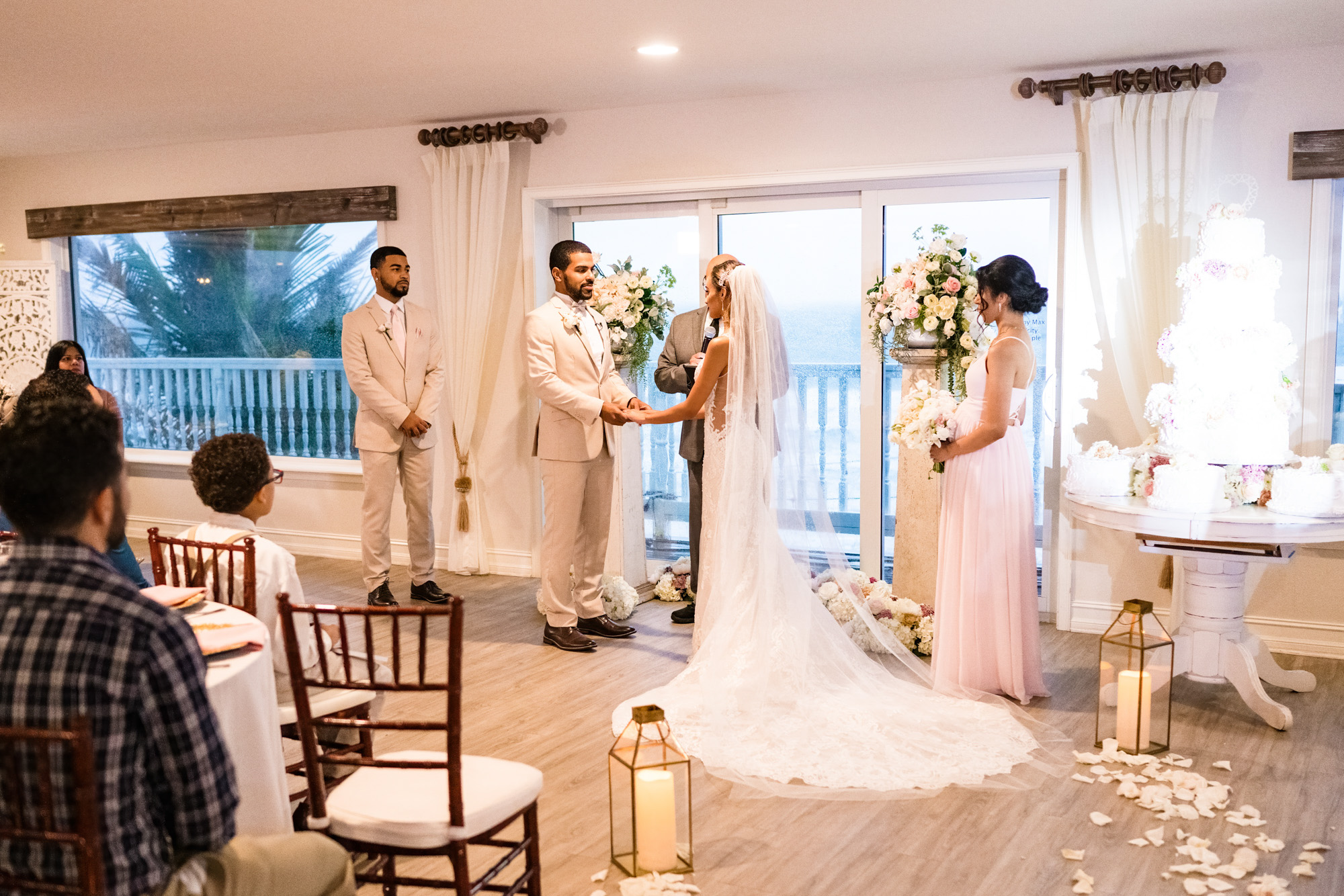 Indoor wedding ceremony moved inside because of rain, with the couple exchanging vows beside floral arrangements and a large wedding cake.