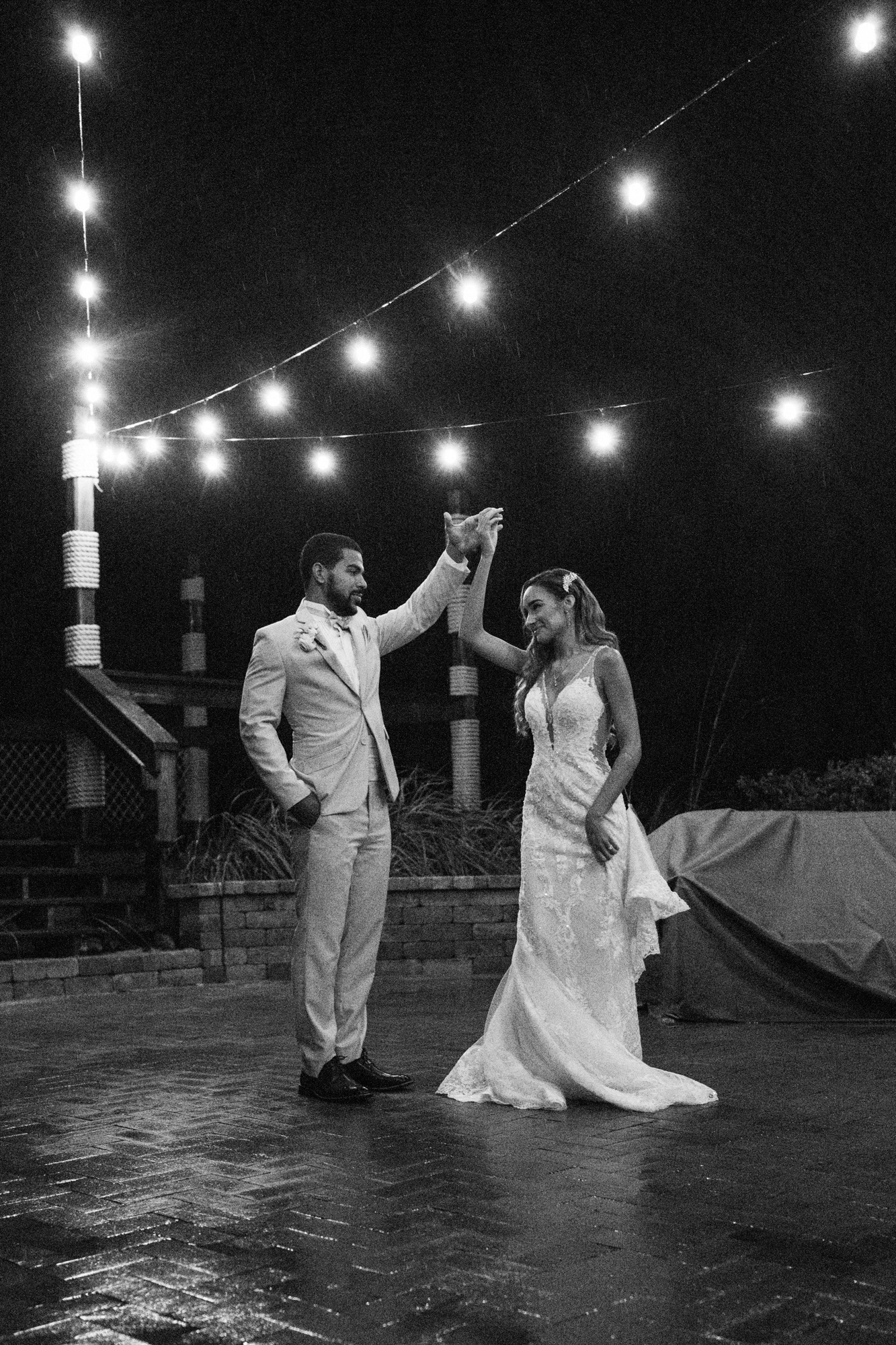 Bride and groom dancing together in the rain beneath string lights after their wedding ceremony.
