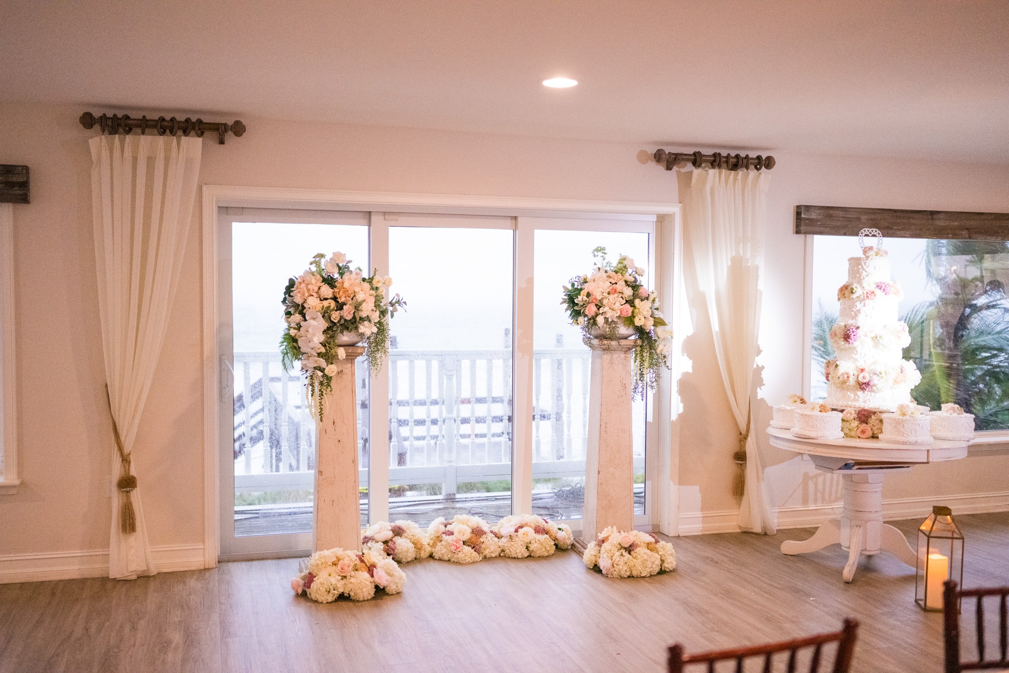 Indoor ceremony setup with floral arrangements, wedding cake, and rain visible through the oceanfront windows.