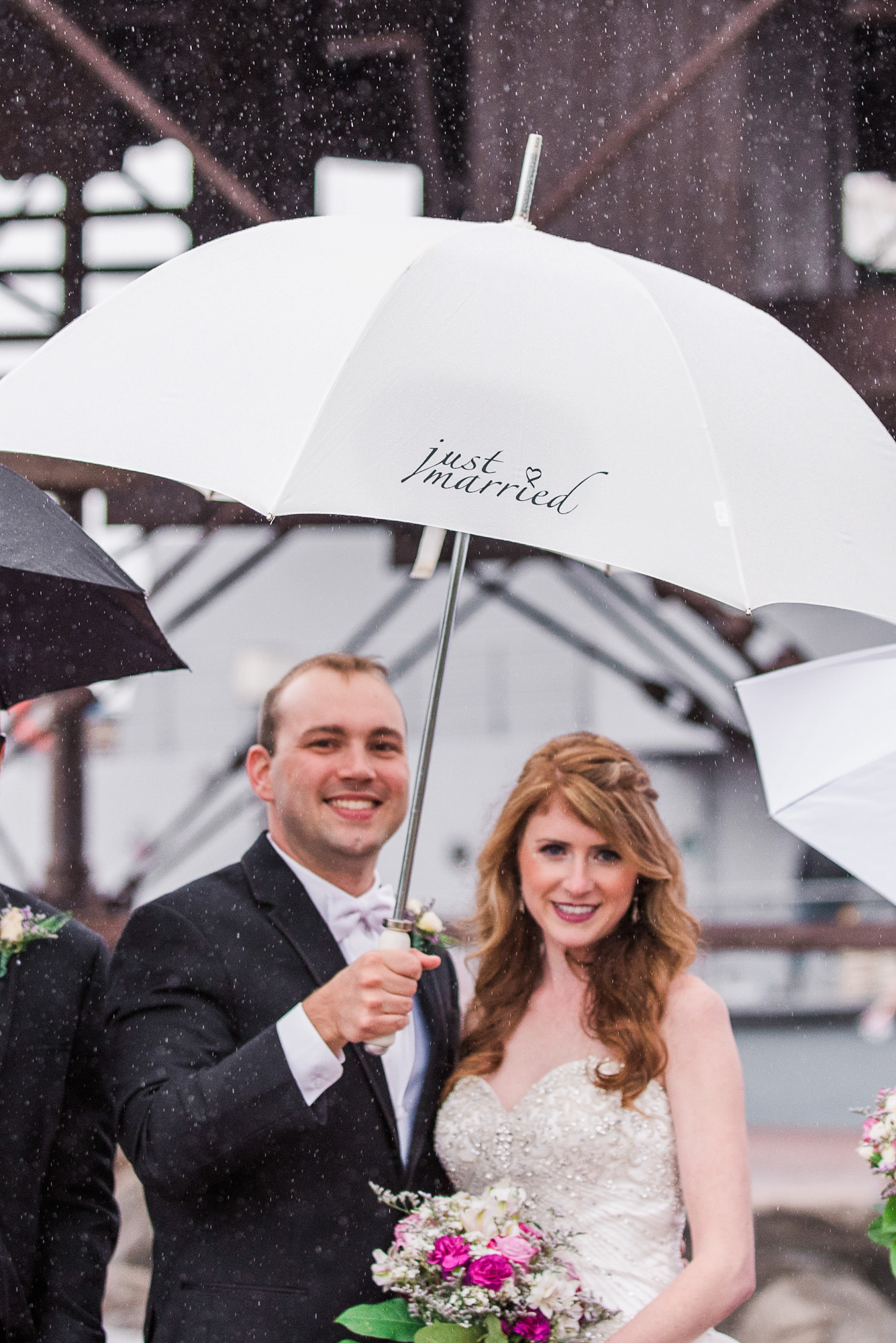 Newlyweds smiling beneath a white “just married” umbrella in the rain.