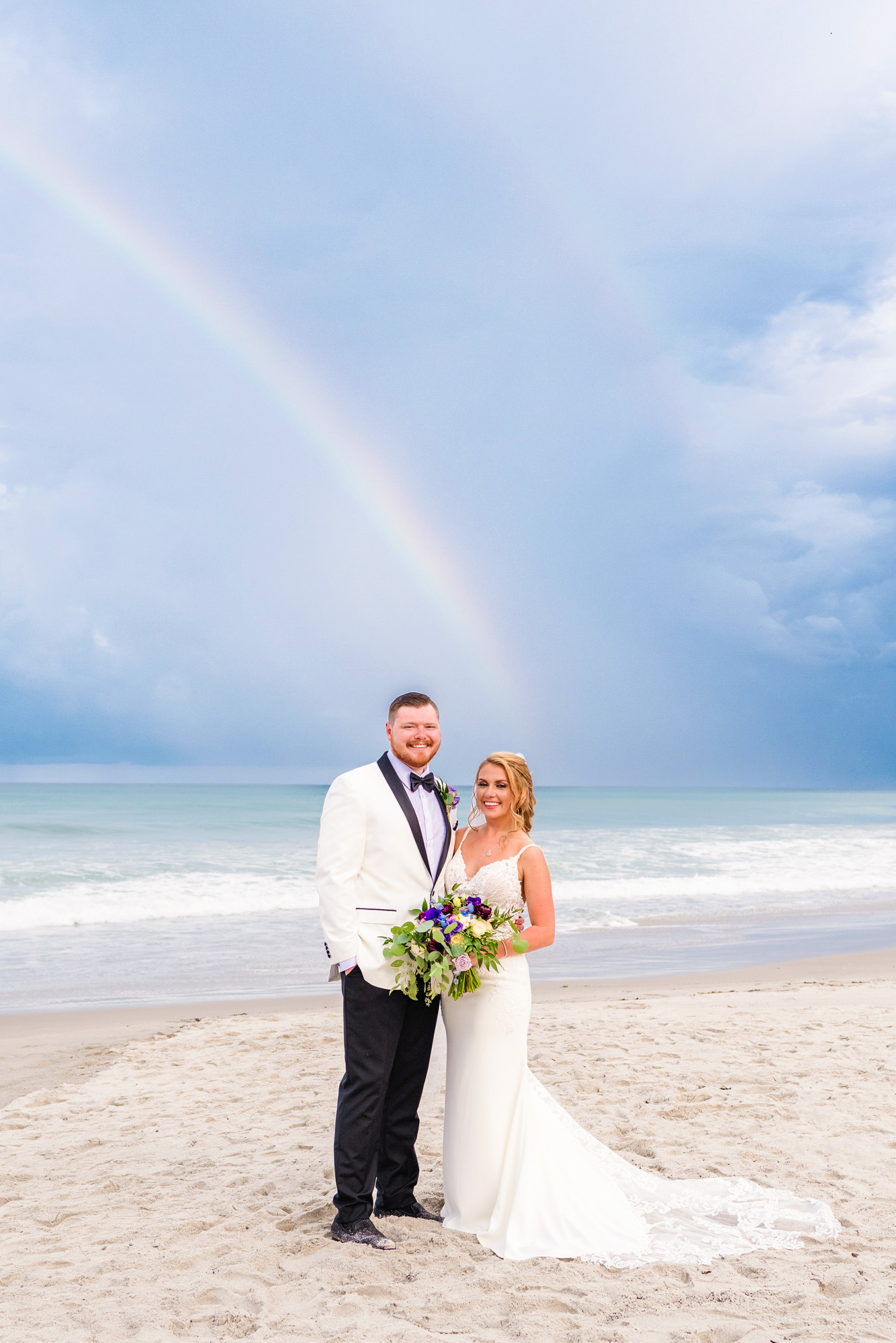 Bride and groom posing on the beach beneath a rainbow after a Florida rain shower at Harbour House Oceanfront.