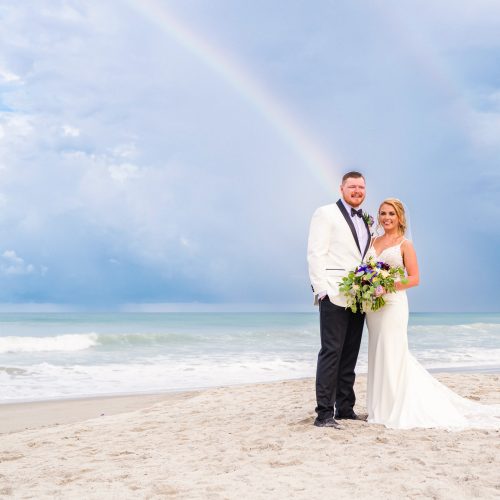 Newlyweds standing on the beach with a rainbow and storm clouds over the ocean.