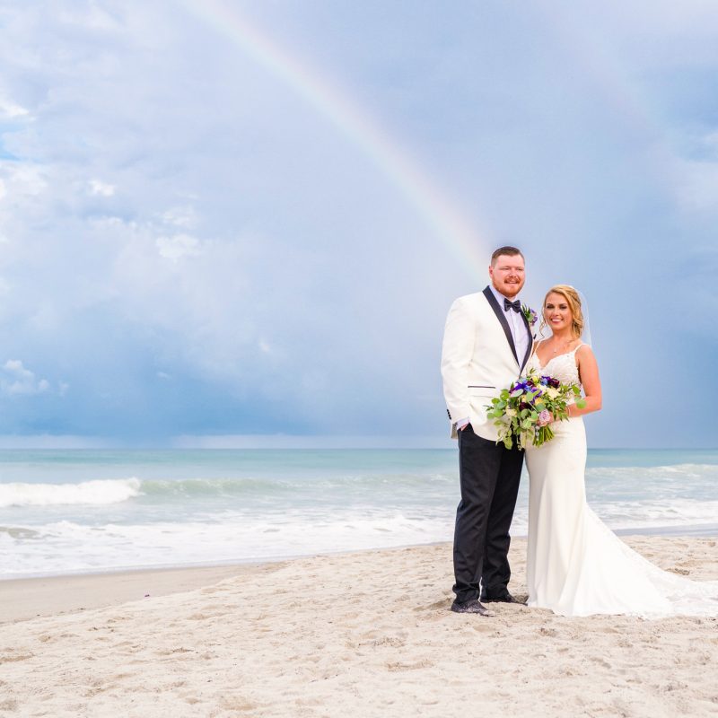 Newlyweds standing on the beach with a rainbow and storm clouds over the ocean.