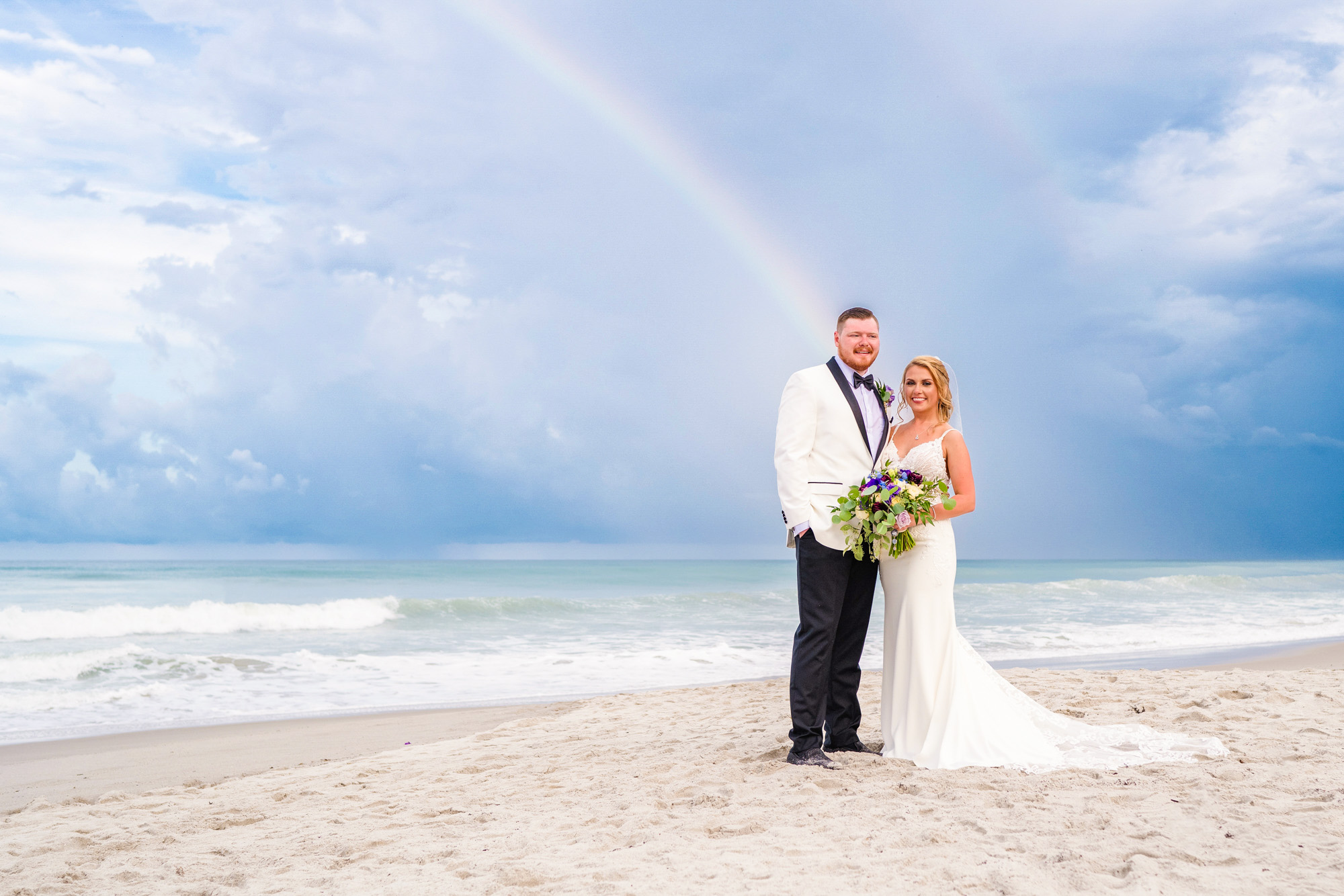 Newlyweds standing on the beach with a rainbow and storm clouds over the ocean.