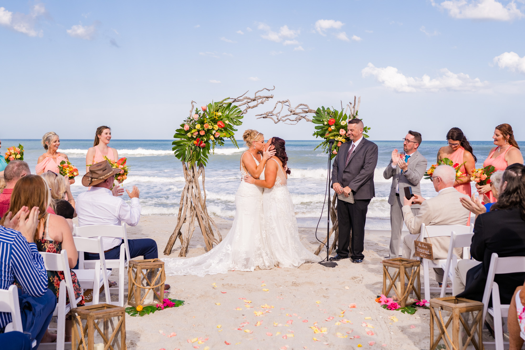 Two brides sharing their first kiss as wedding guests applaud on the beach.