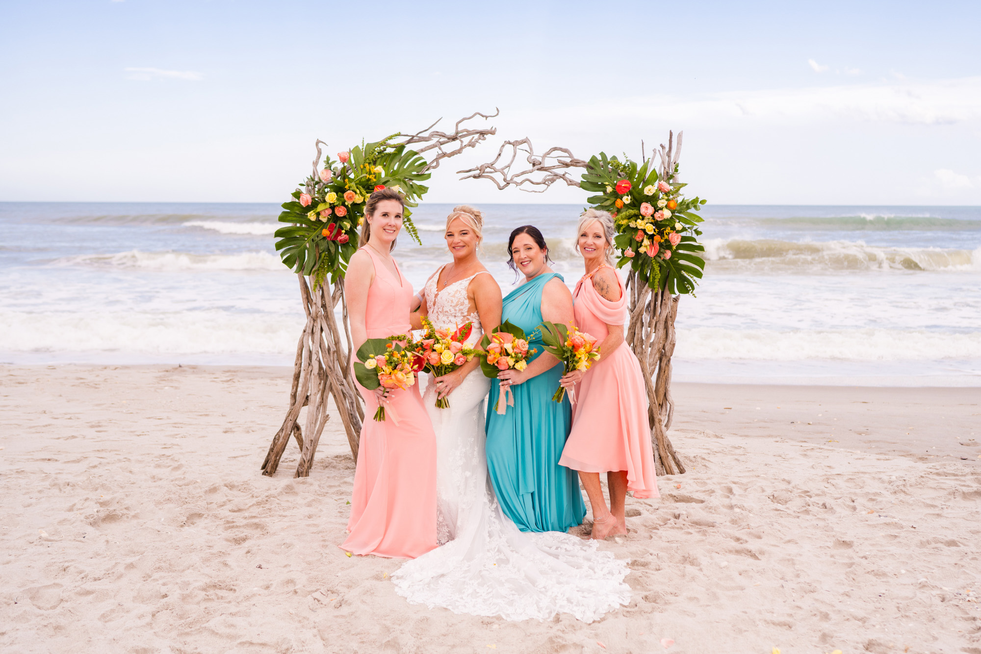 Bride posing with members of her wedding party beneath a tropical beach wedding arbor.