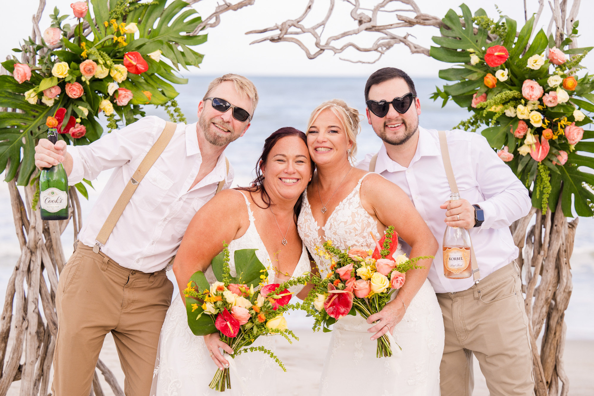 Two brides smiling with members of their wedding party beneath a tropical floral arbor.