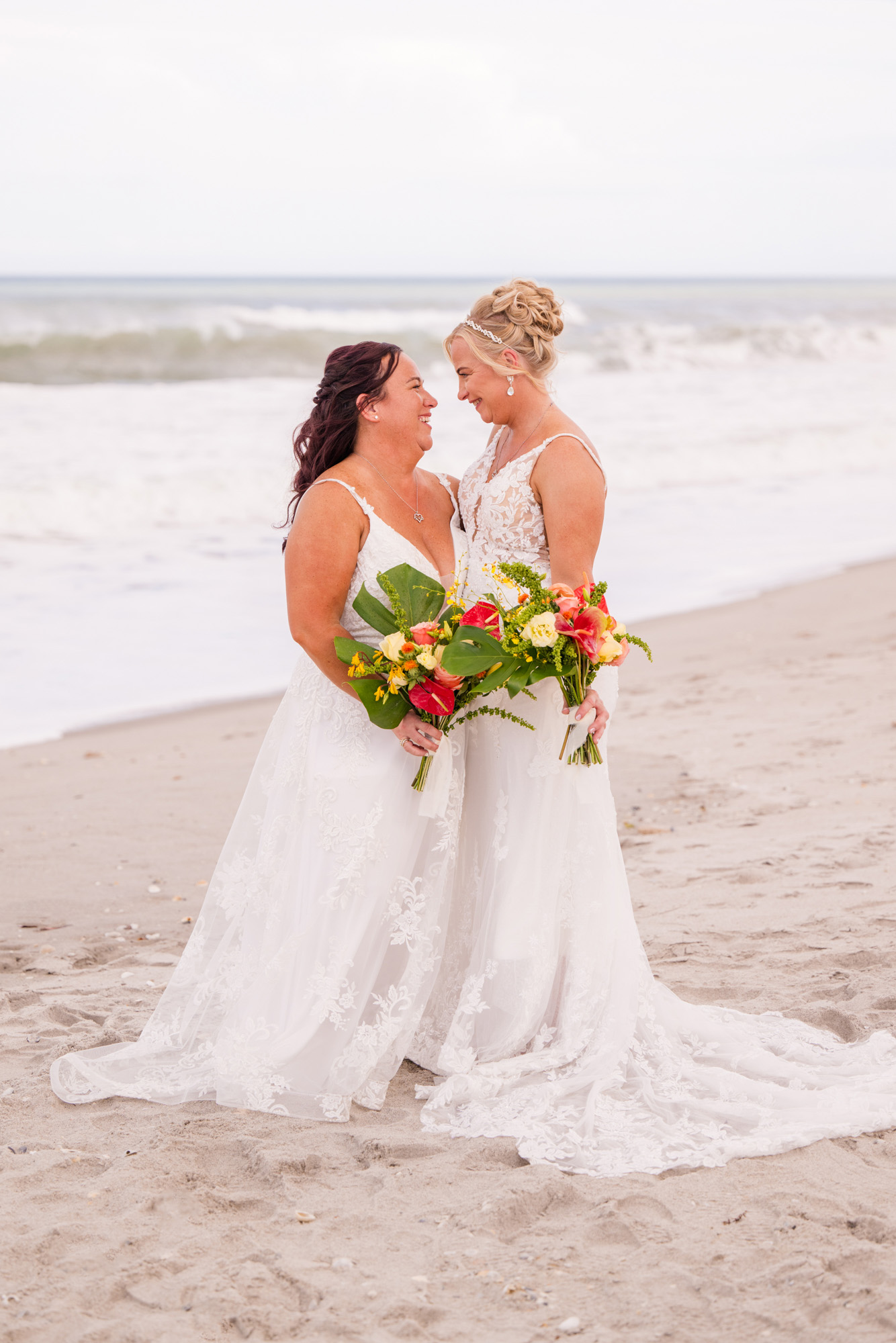 Two brides smiling at each other while holding colorful tropical bouquets on the beach.