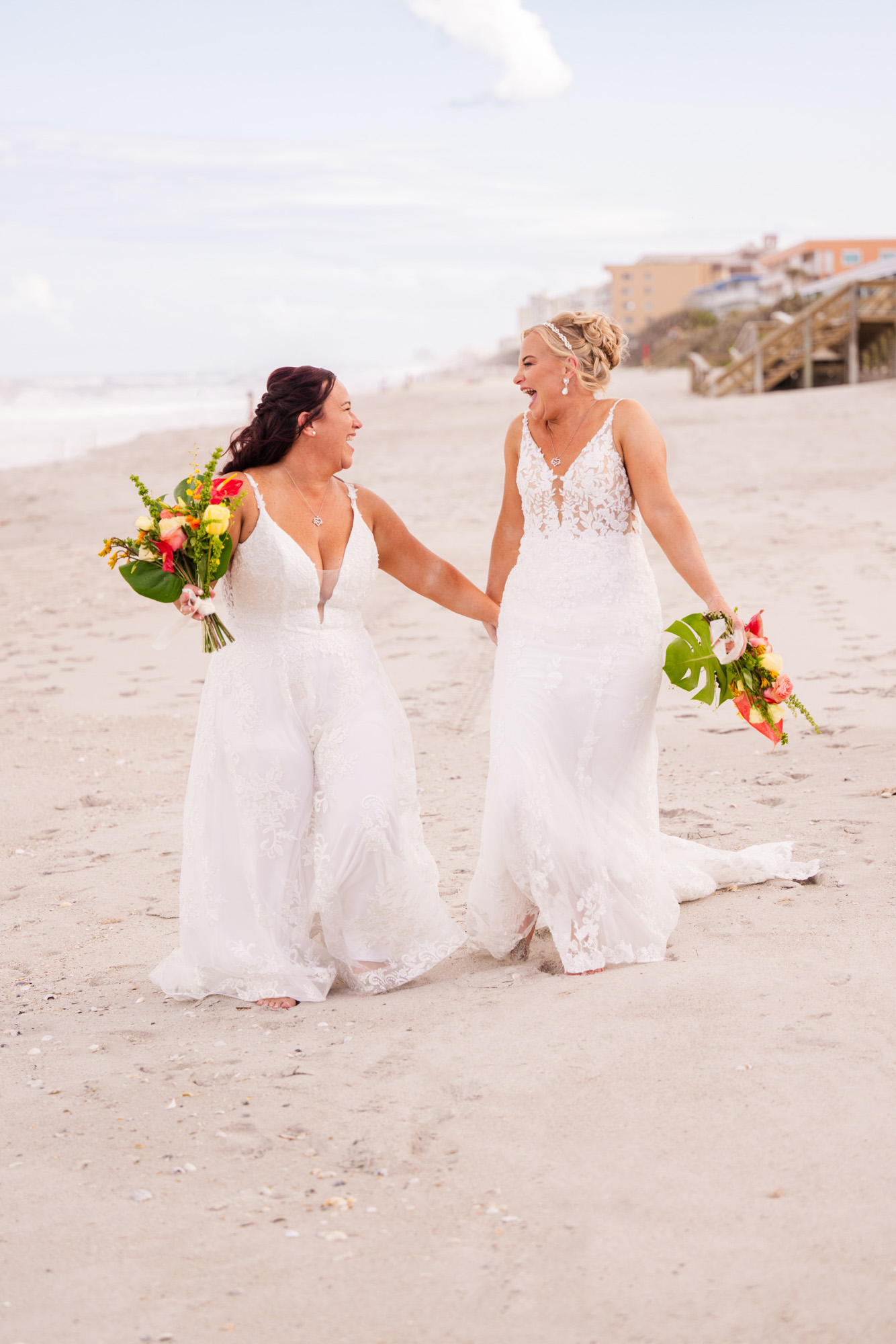 Two brides laughing together while walking hand in hand along the beach with their bouquets.