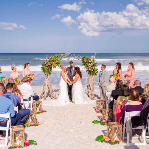 Wide oceanfront wedding ceremony with two brides standing beneath a tropical floral arbor.
