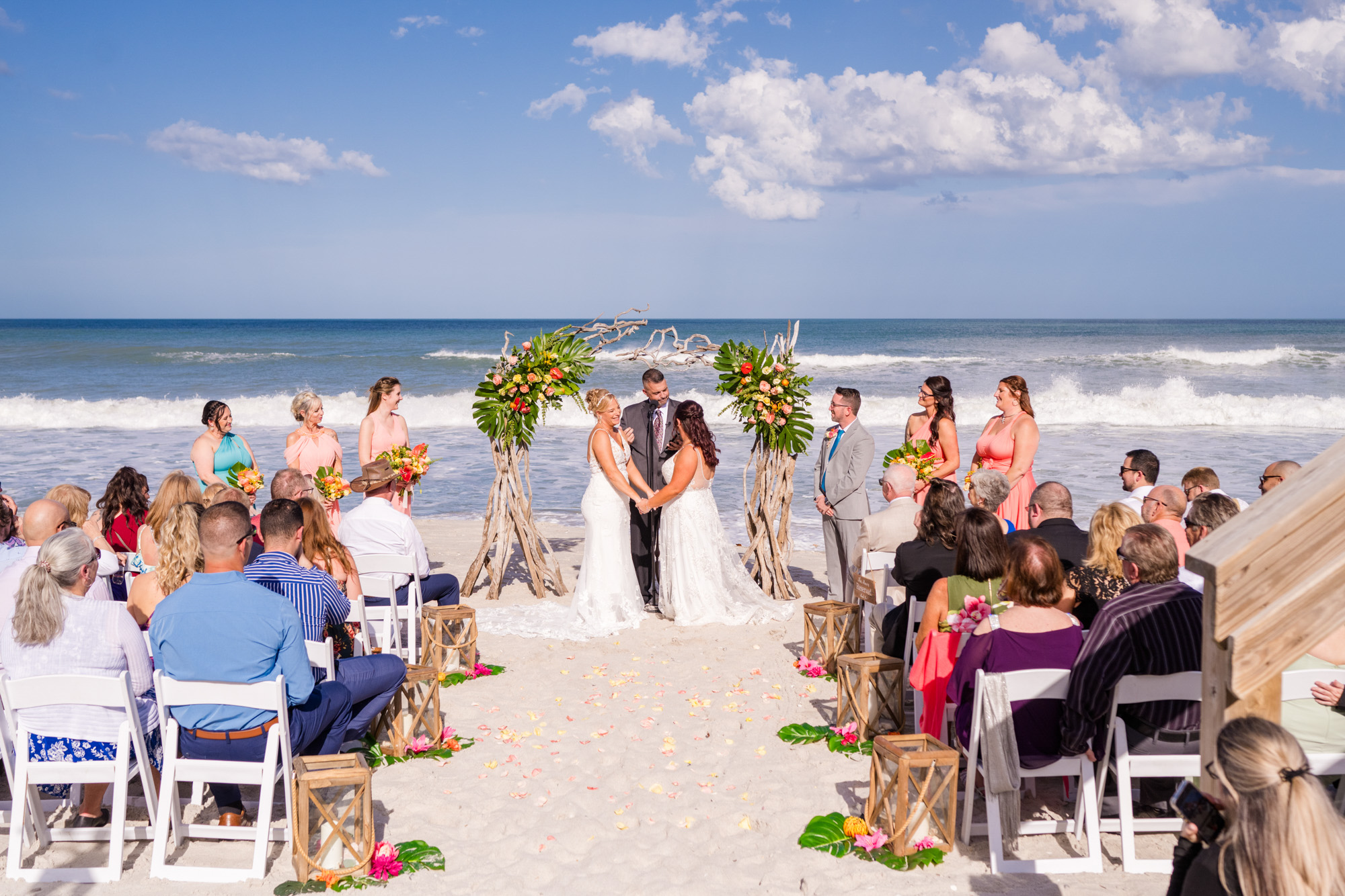 Wide oceanfront wedding ceremony with two brides standing beneath a tropical floral arbor.