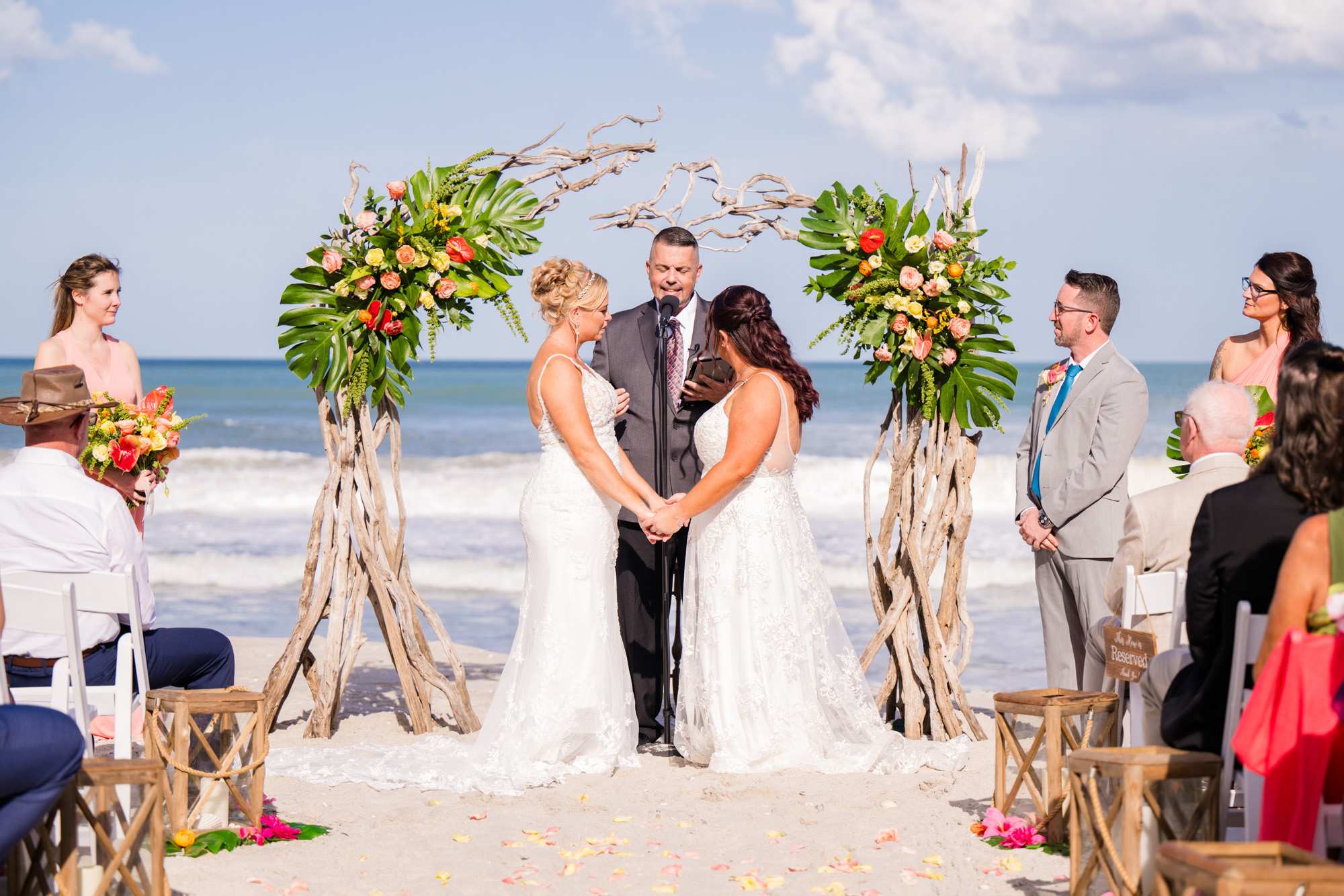 Two brides holding hands at their beach wedding ceremony framed by a driftwood and tropical flower arbor.