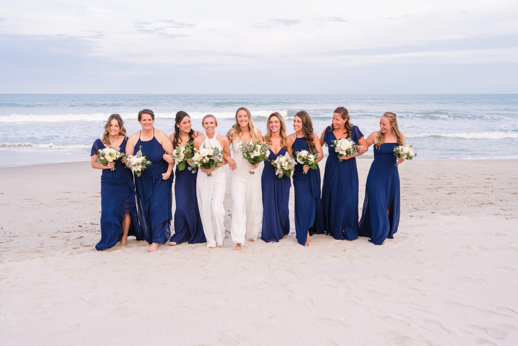 Two brides walking with their bridesmaids along the beach carrying white and green bouquets.