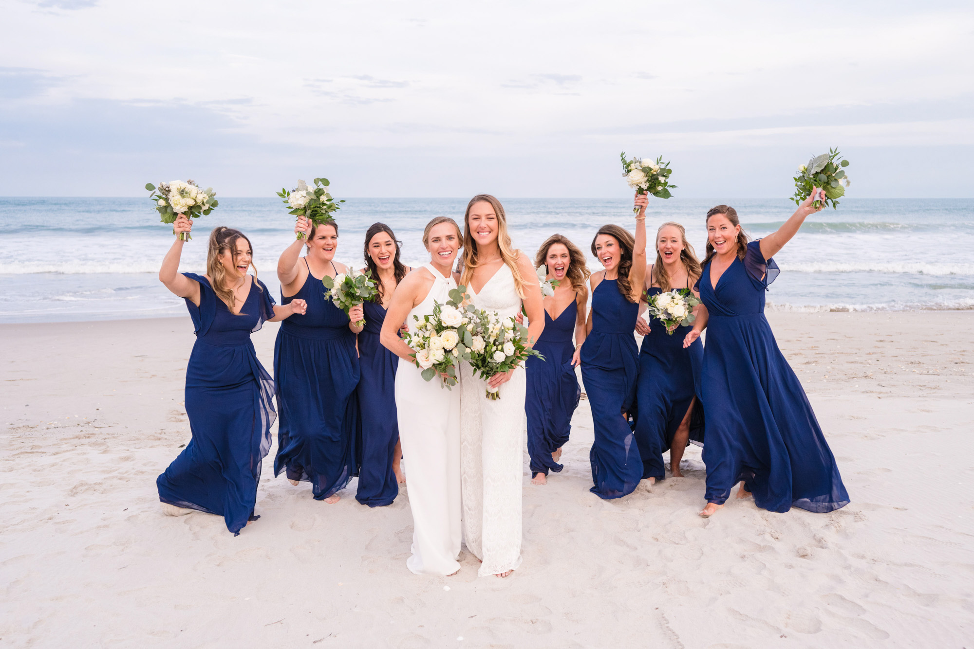 Two brides posing with their bridesmaids in navy dresses during oceanfront wedding portraits.
