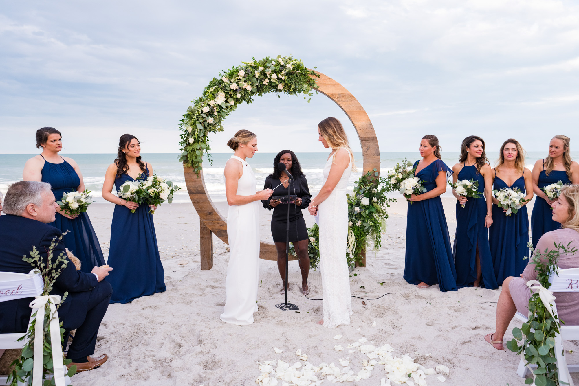 Two brides standing hand in hand during their oceanfront ceremony with bridesmaids in navy dresses.