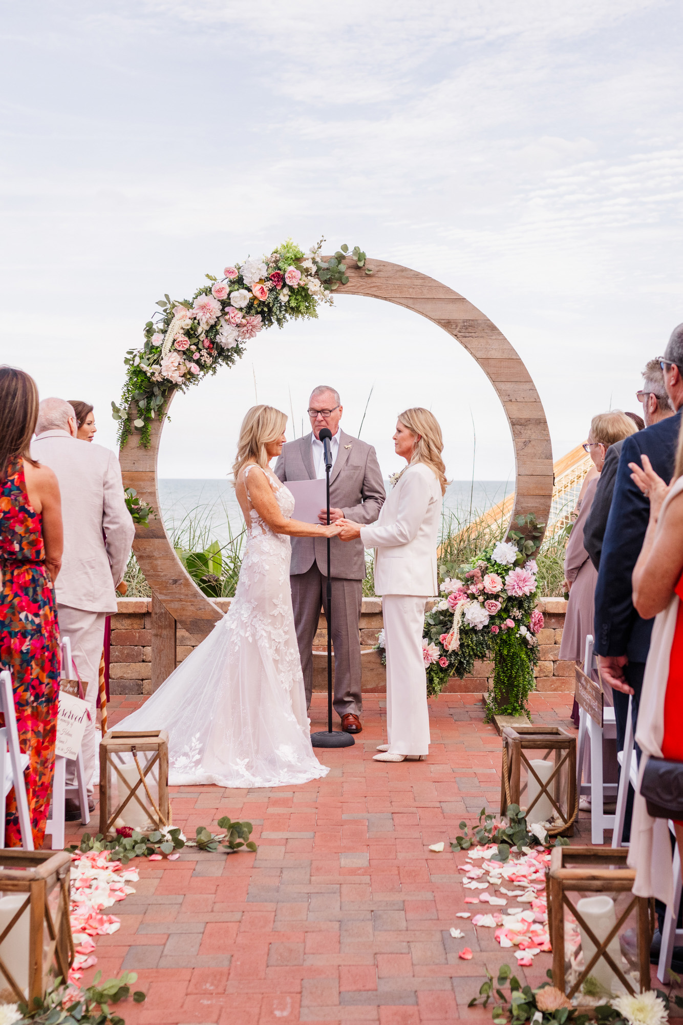 Wide view of two brides exchanging vows beneath a large circular flower-covered wedding arbor.