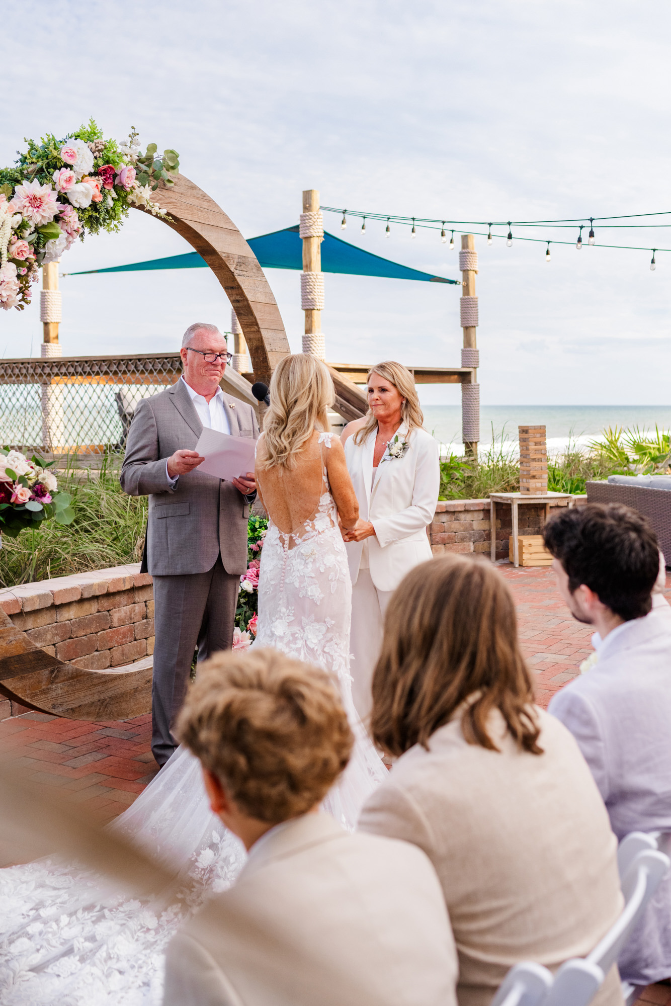 Two brides exchanging vows during an oceanfront ceremony with guests seated nearby.
