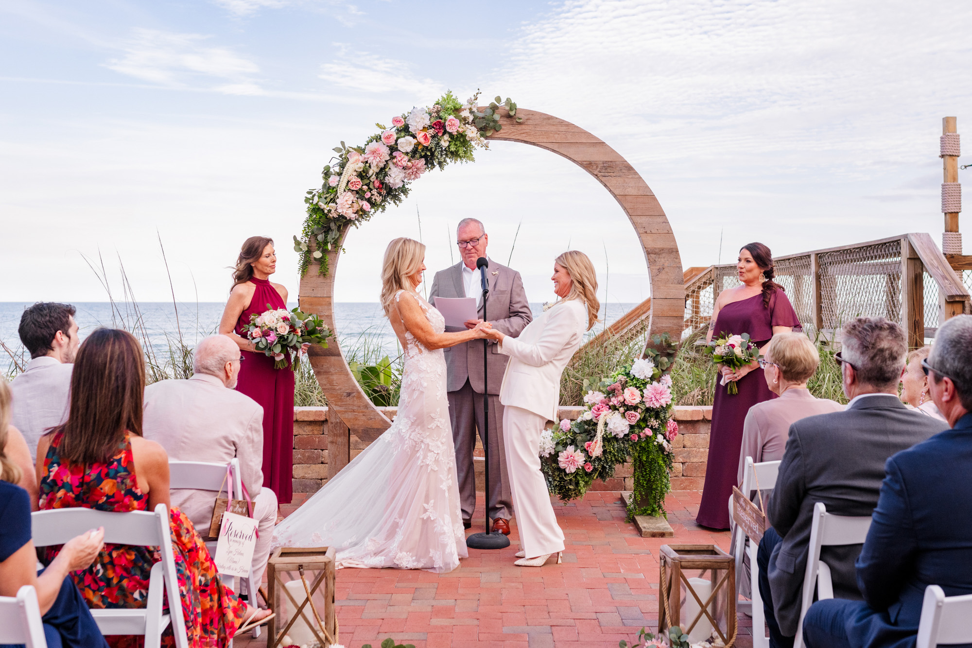 Wide view of a same-sex oceanfront wedding ceremony beneath a circular pink floral arbor.
