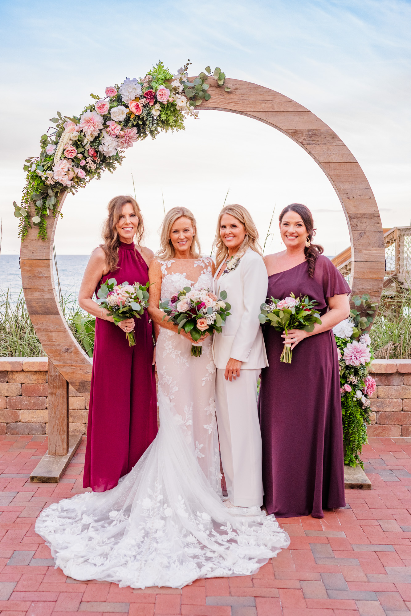 Two brides posing with their wedding attendants beneath a circular floral arbor by the ocean.