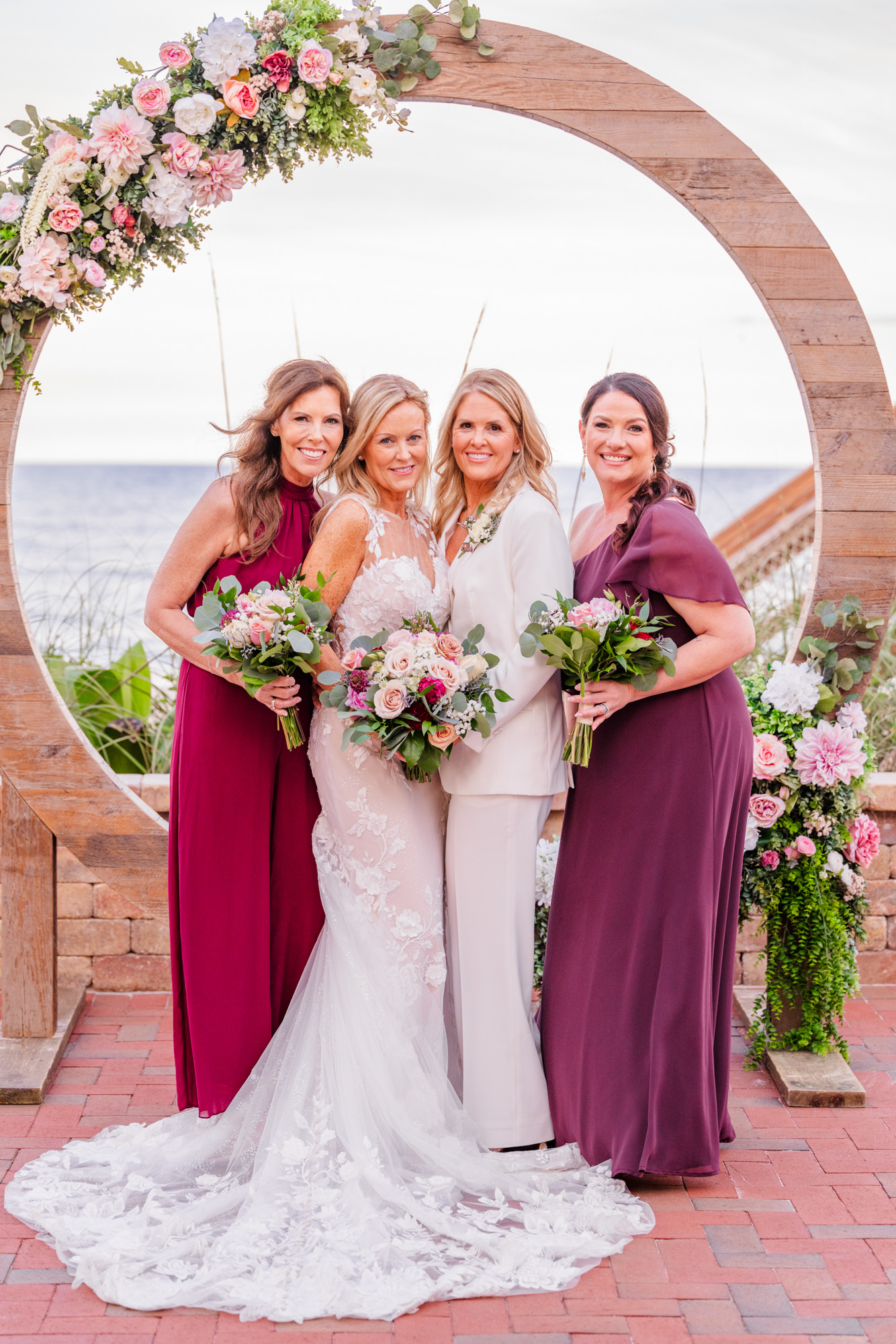 Two brides standing close together with bridesmaids in burgundy dresses at an oceanfront wedding.