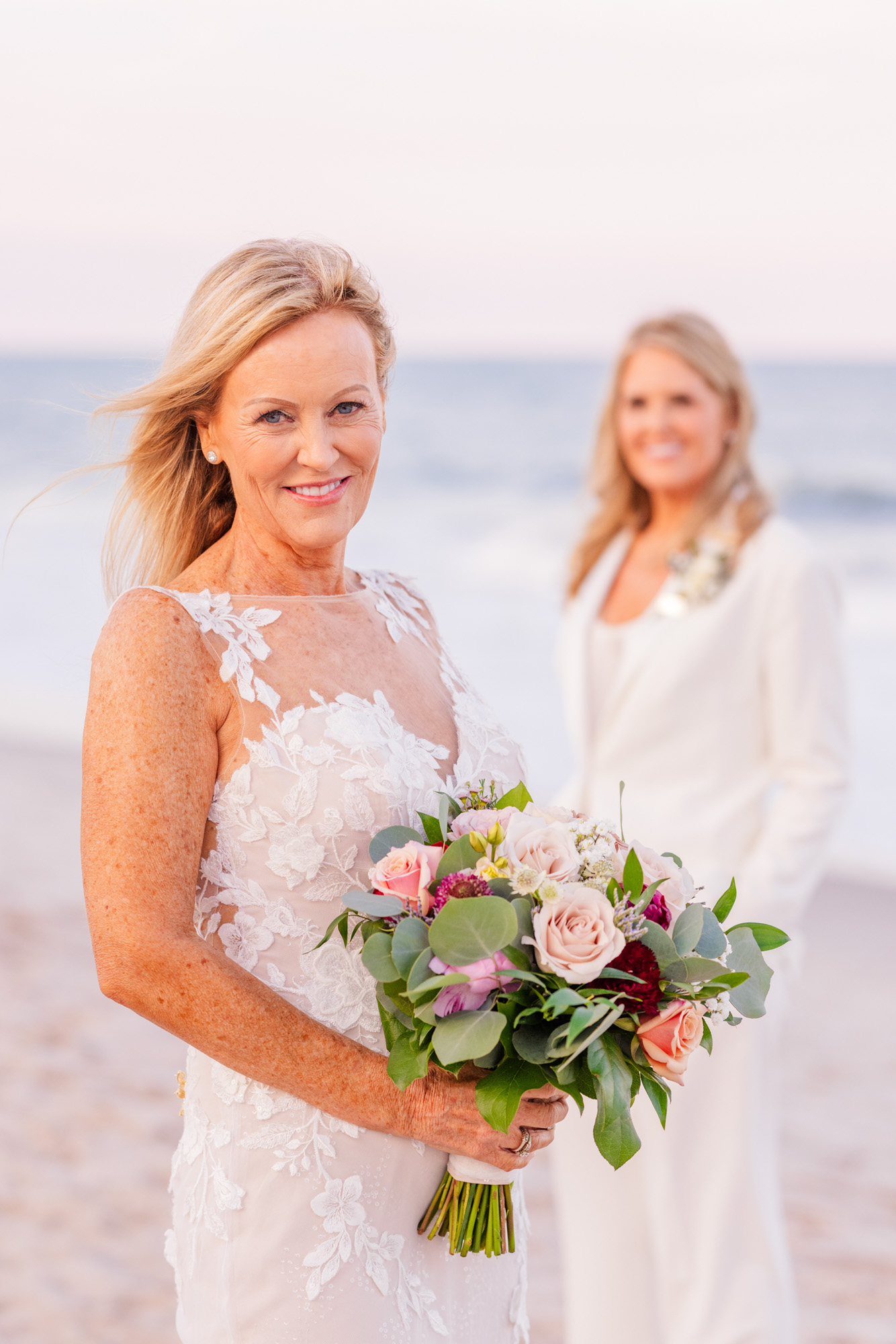 Bride in a white suit smiling toward her partner during beach portraits.