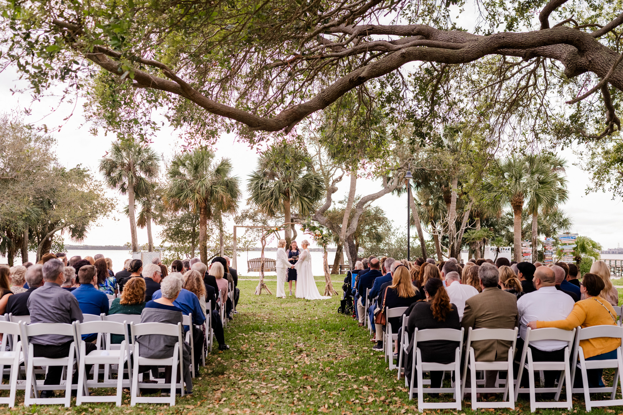 Wide view of a same-sex wedding ceremony under large oak trees with guests seated on white chairs.
