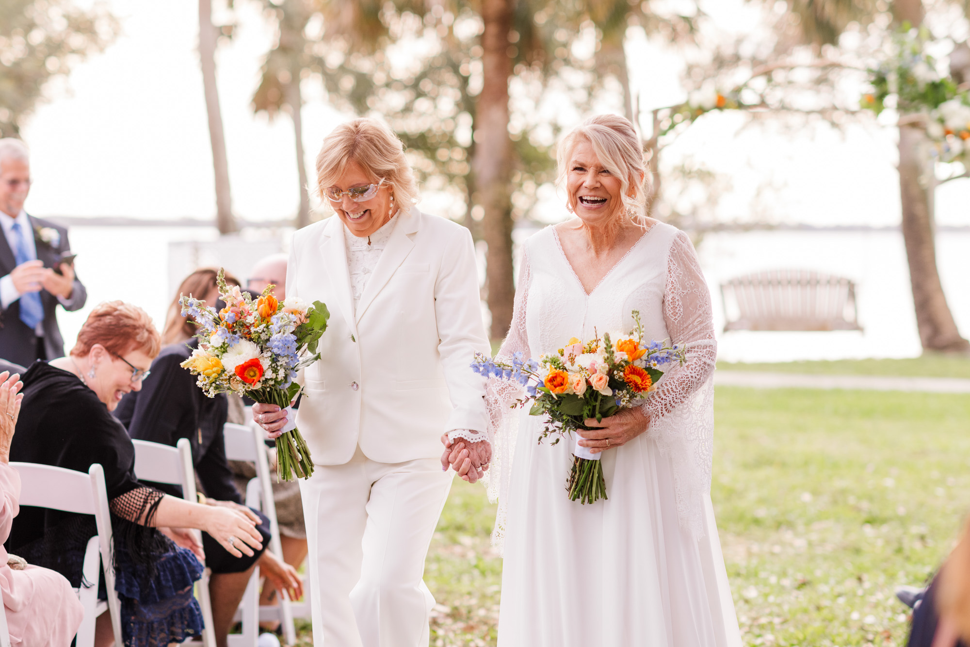 Two brides laughing together while carrying colorful wedding bouquets after their ceremony.