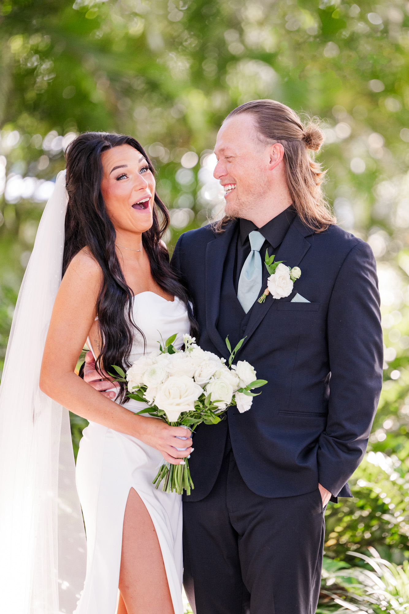 Bride laughing with her groom while holding a white wedding bouquet outdoors.