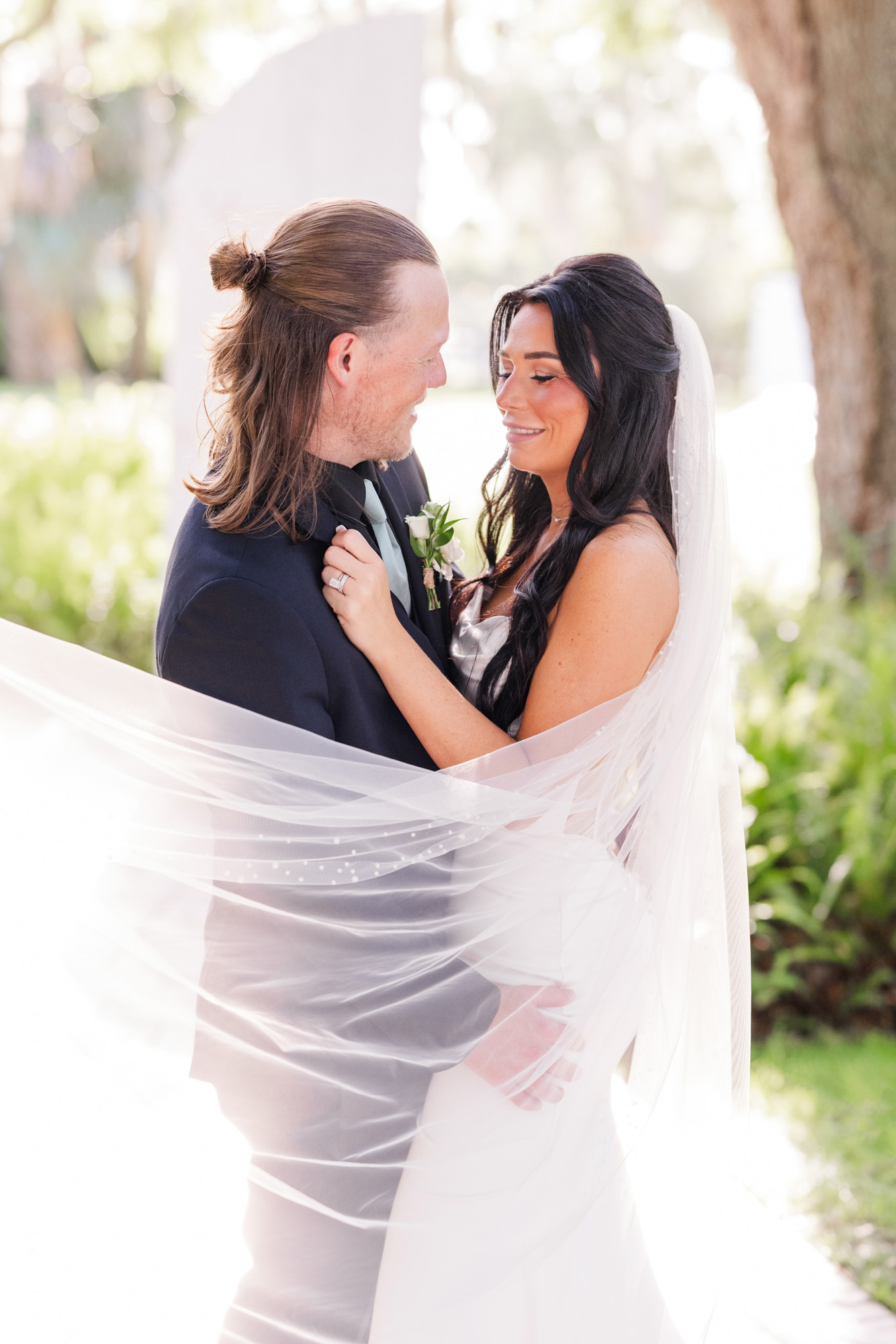 Bride and groom standing close together while her veil wraps around them during portraits.