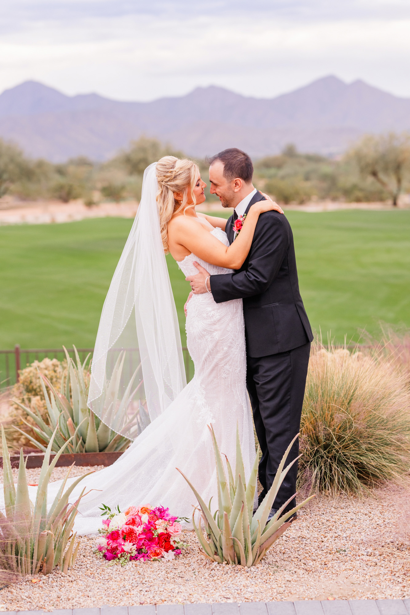 Bride and groom standing together during romantic wedding portraits beside desert landscaping and mountains.