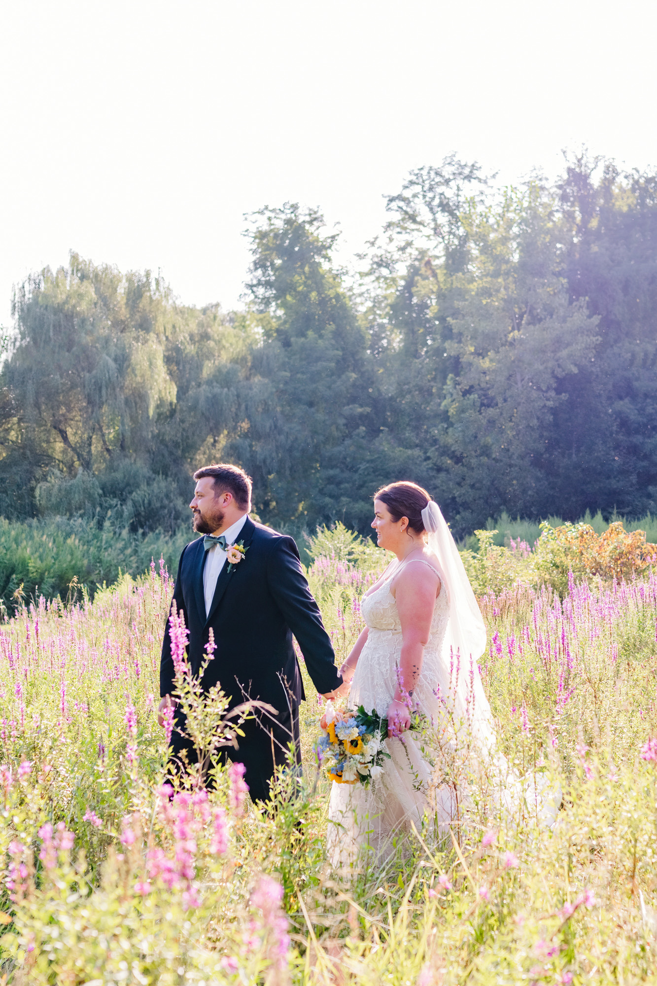 Bride and groom holding hands while walking through a sunlit wildflower field.