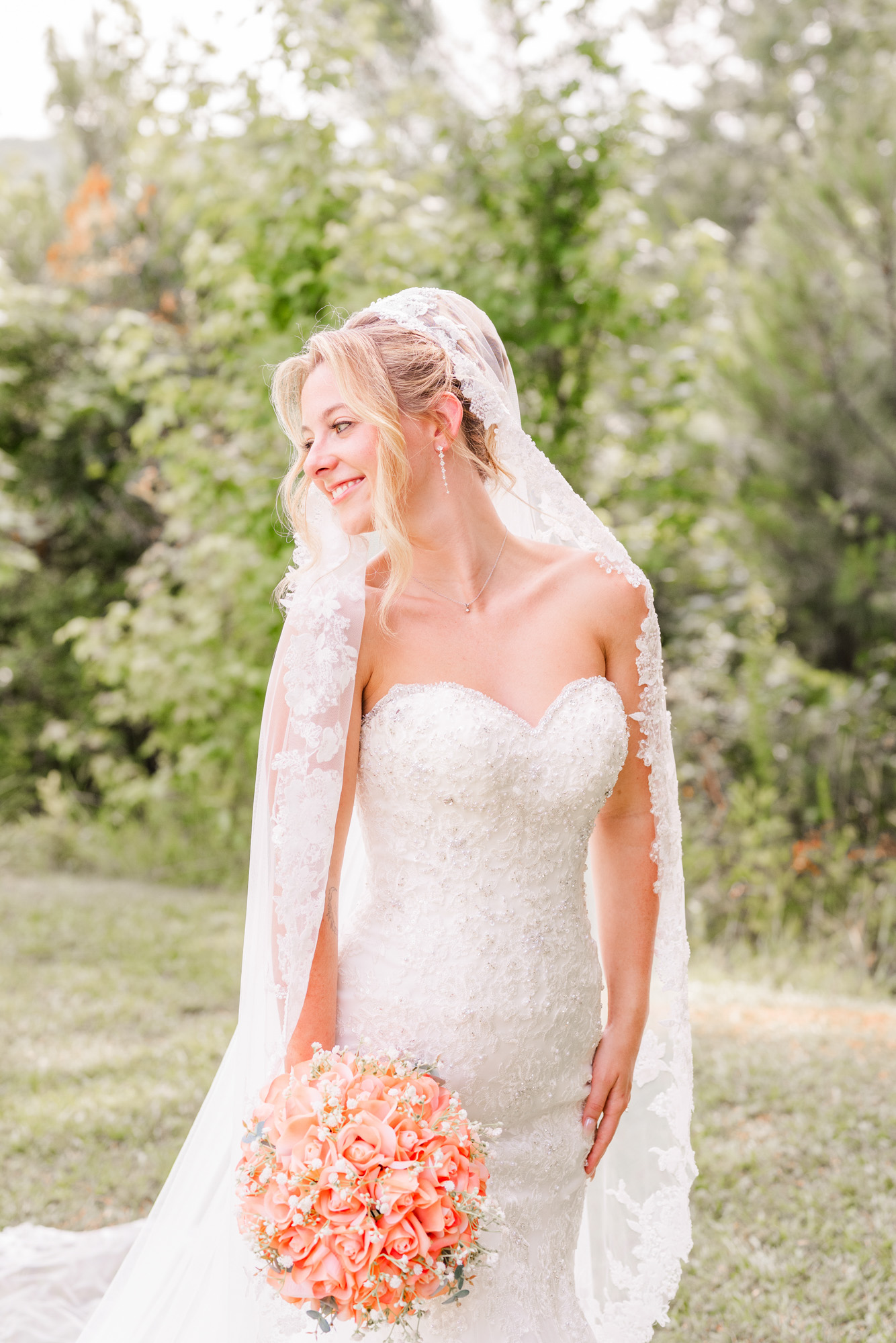 Bride posing outdoors in a lace wedding gown and veil with a coral bouquet.
