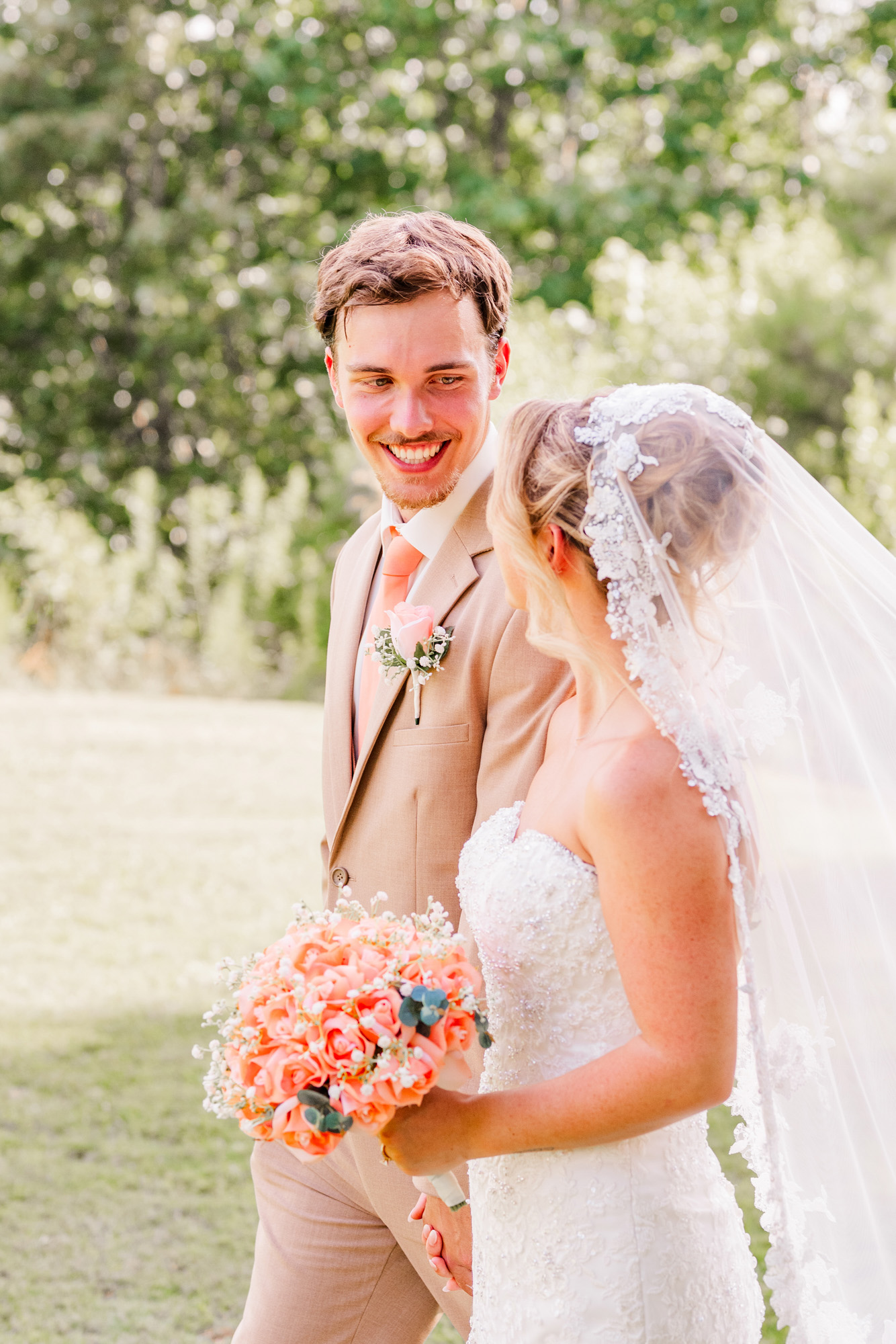 Groom smiling at the bride as she holds a coral wedding bouquet.