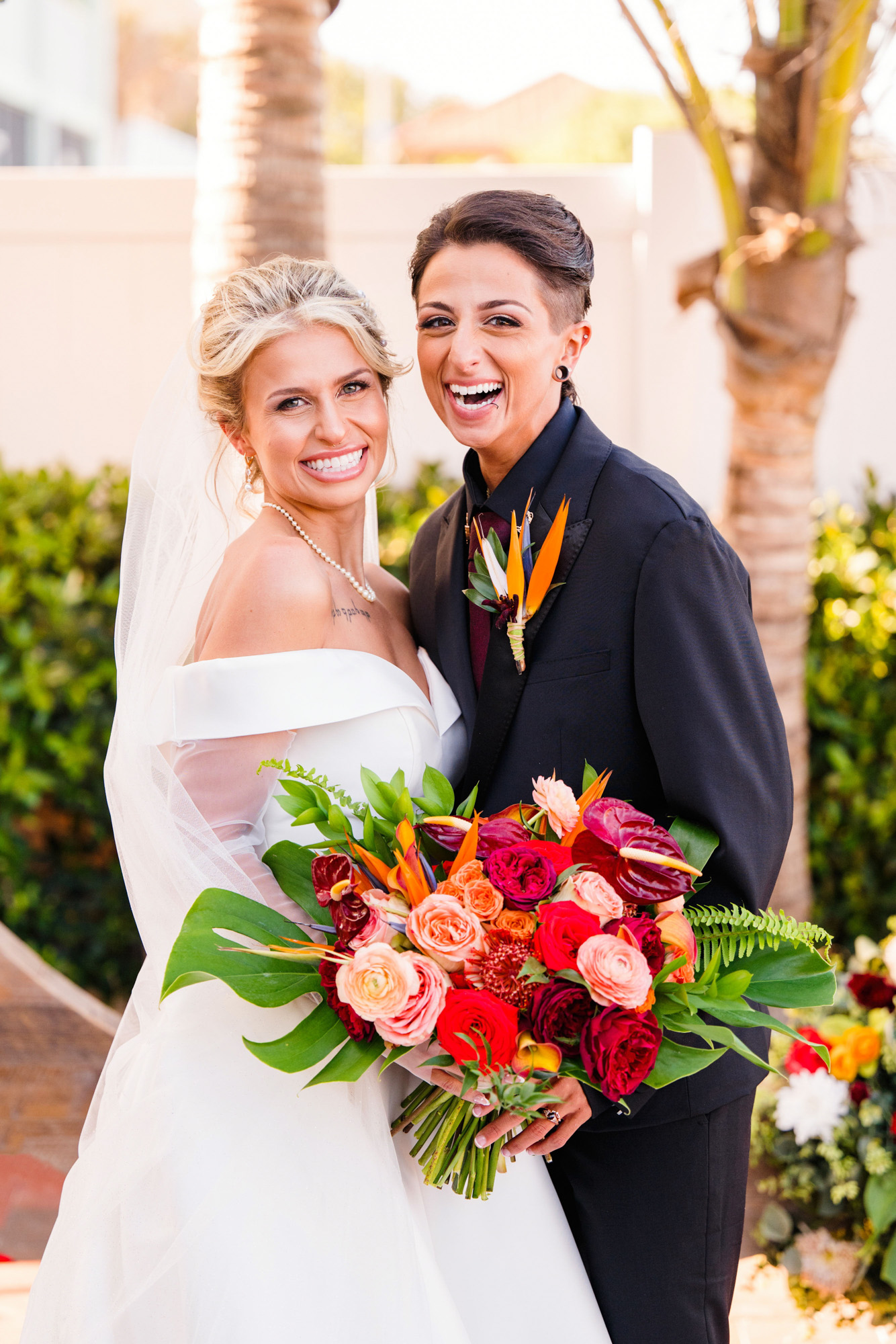 Two brides laughing together while holding a vibrant tropical bouquet.