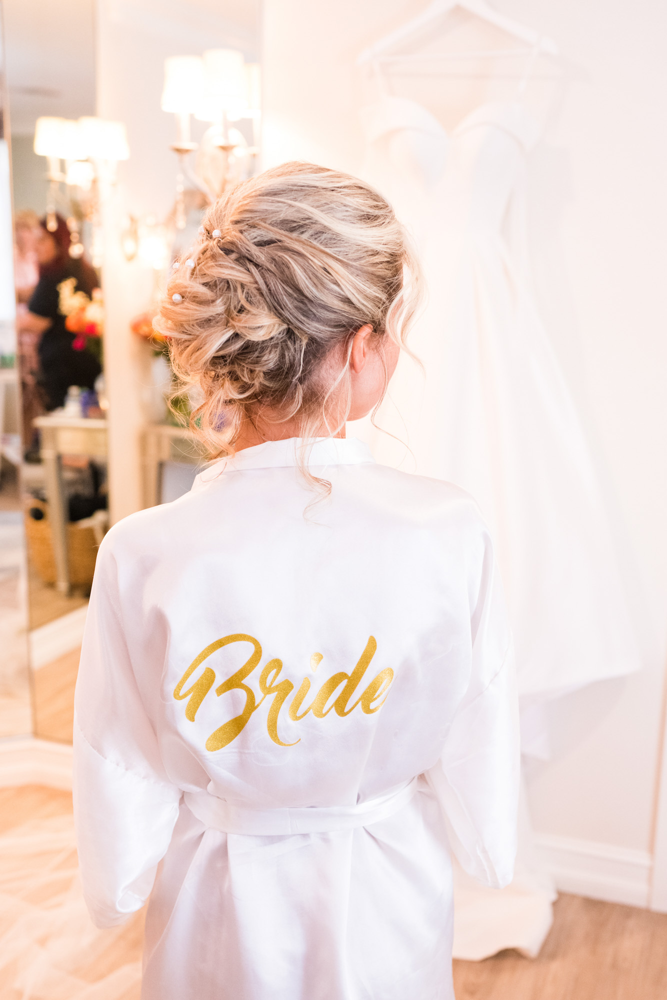 Bride in a white satin robe with “Bride” written in gold on the back, standing near her wedding dress.