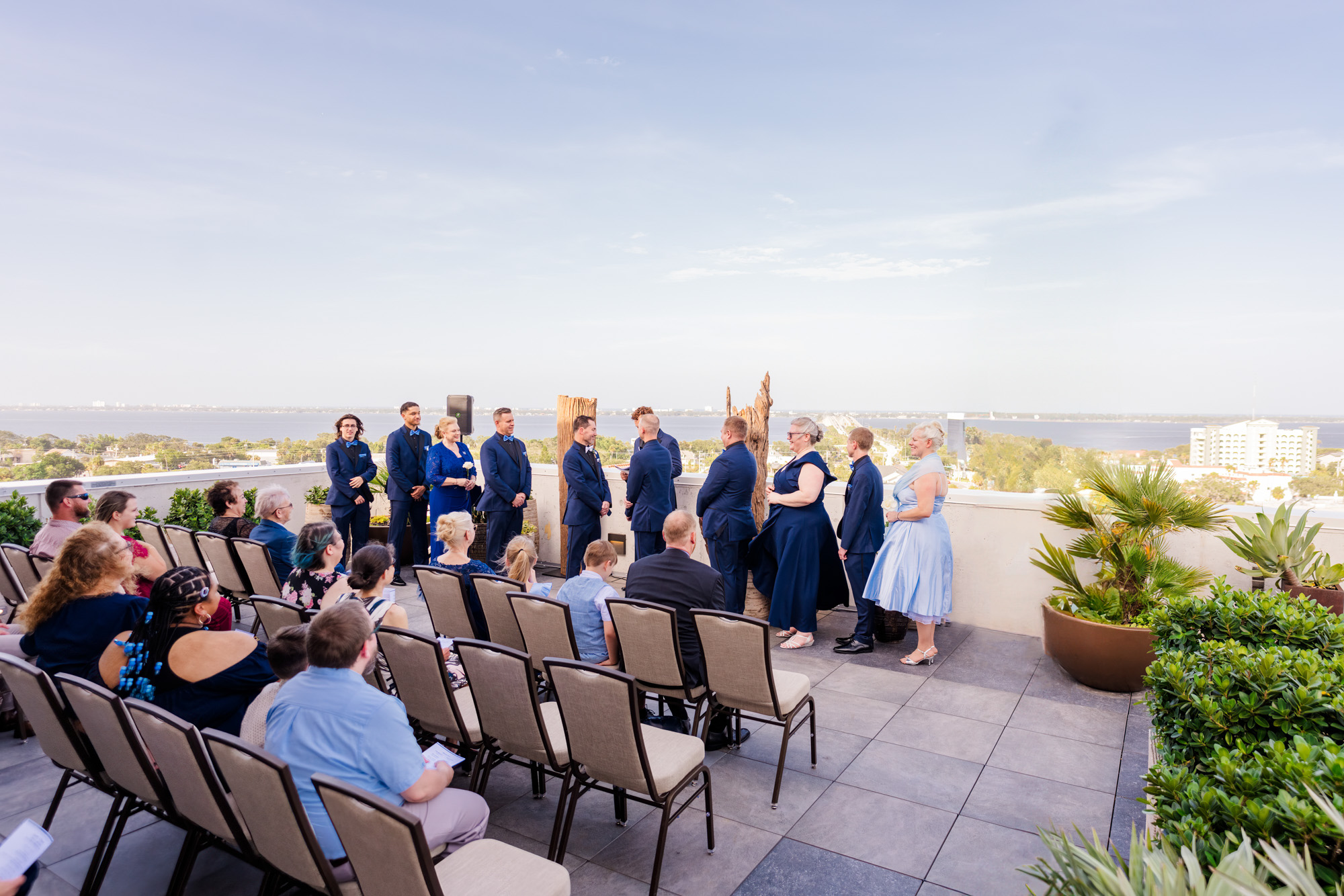 Two grooms facing each other during their rooftop wedding ceremony with attendants nearby.