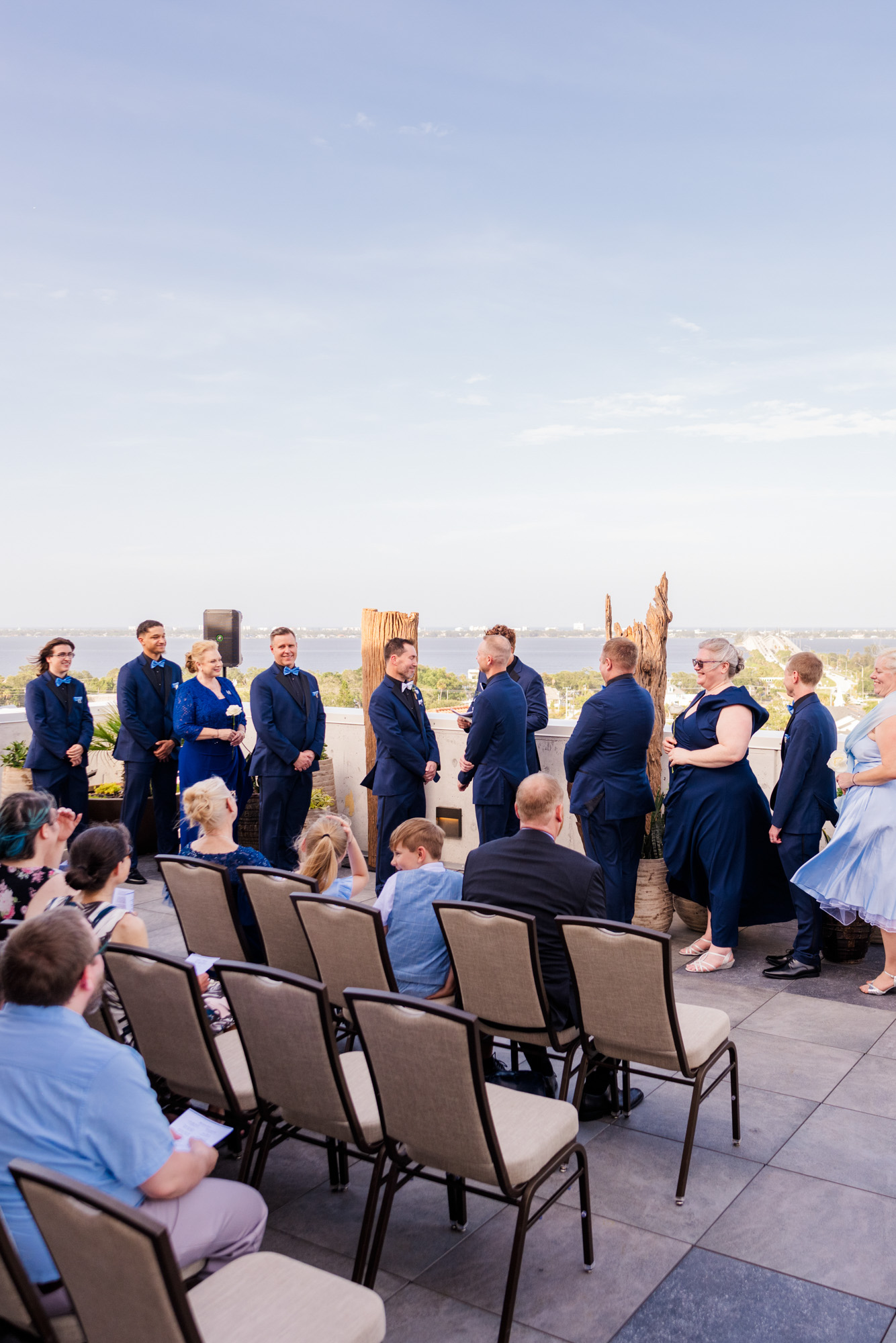 Wide view of a same-sex rooftop wedding ceremony with guests seated and a city-and-water view beyond.