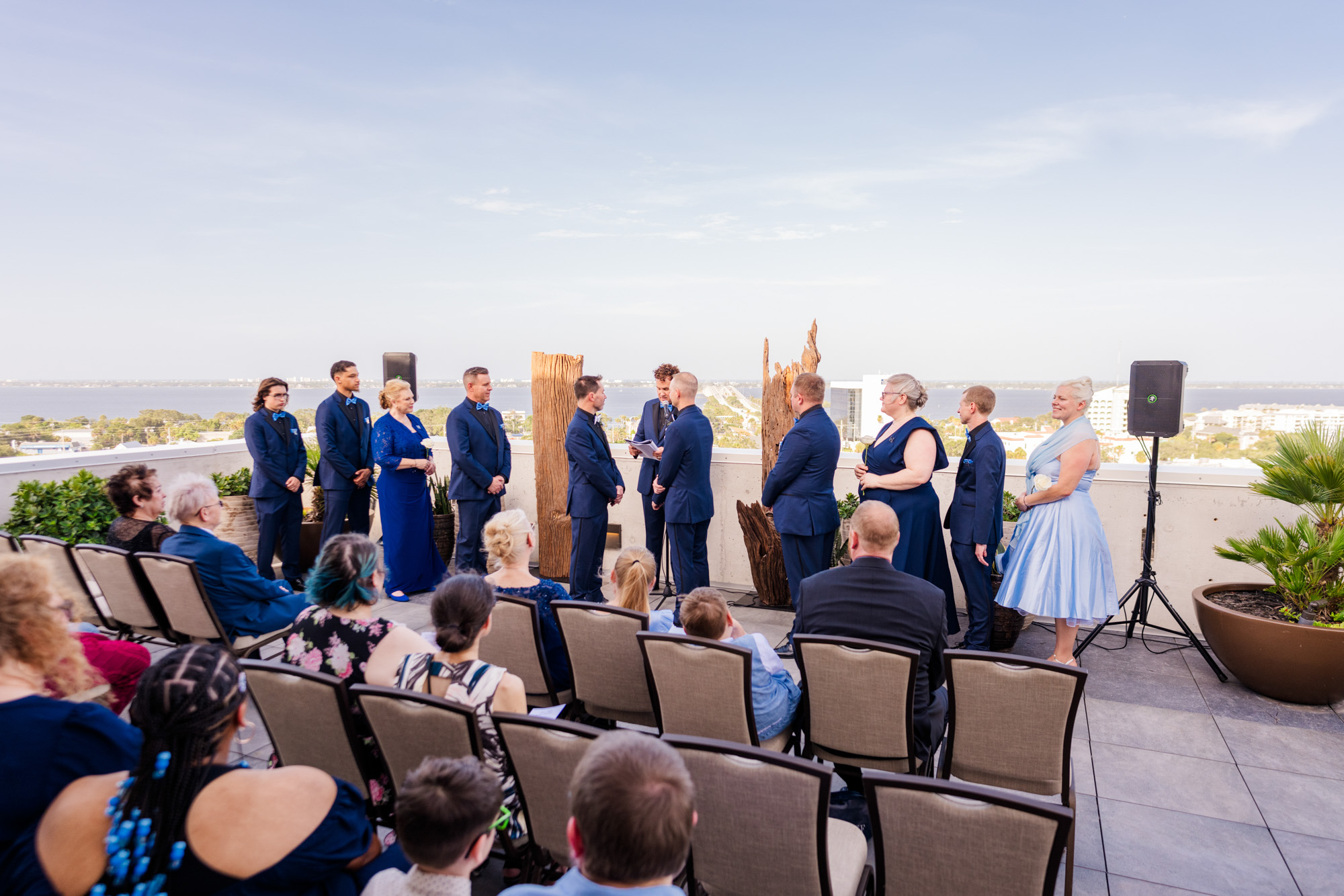 Wide rooftop wedding ceremony with two grooms, wedding party, and guests overlooking the water at Hotel Melby in Melbourne, Florida