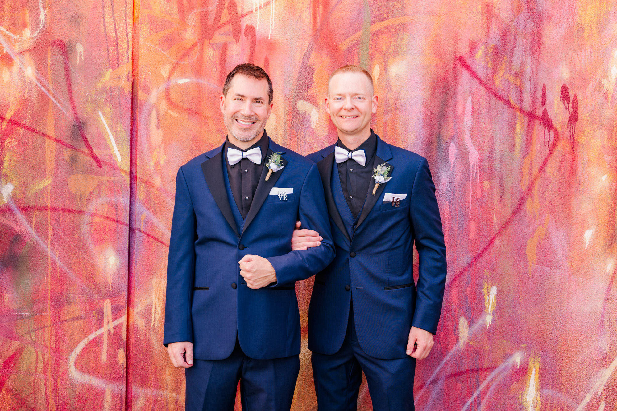 Two grooms posing arm in arm in front of a colorful mural at their Hotel Melby wedding.
