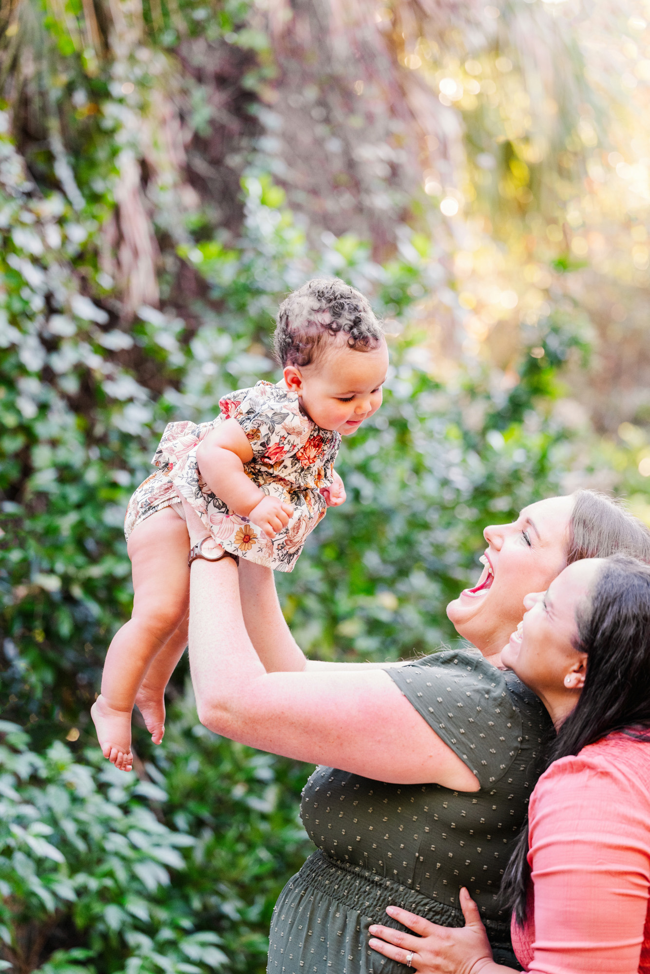 Mother smiling up at her baby as she lifts the child beneath the trees.