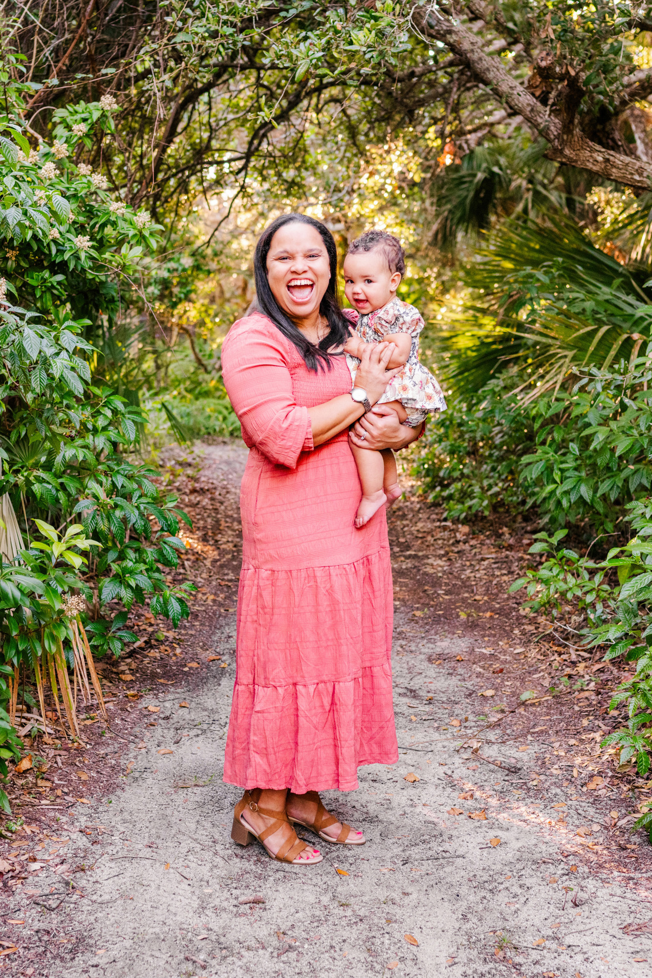 Mother in a coral dress holding her baby on a shaded garden path.