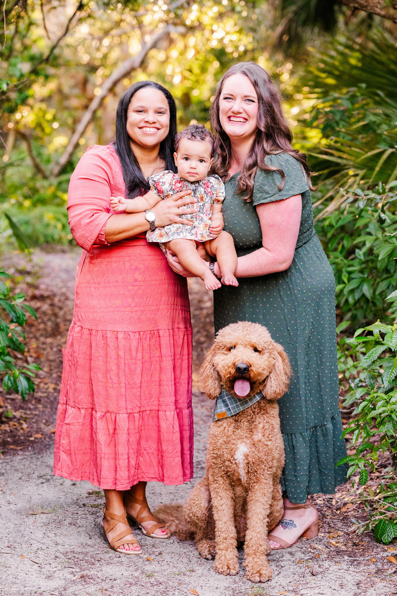 Two moms holding their baby beside their dog during a colorful outdoor family portrait.