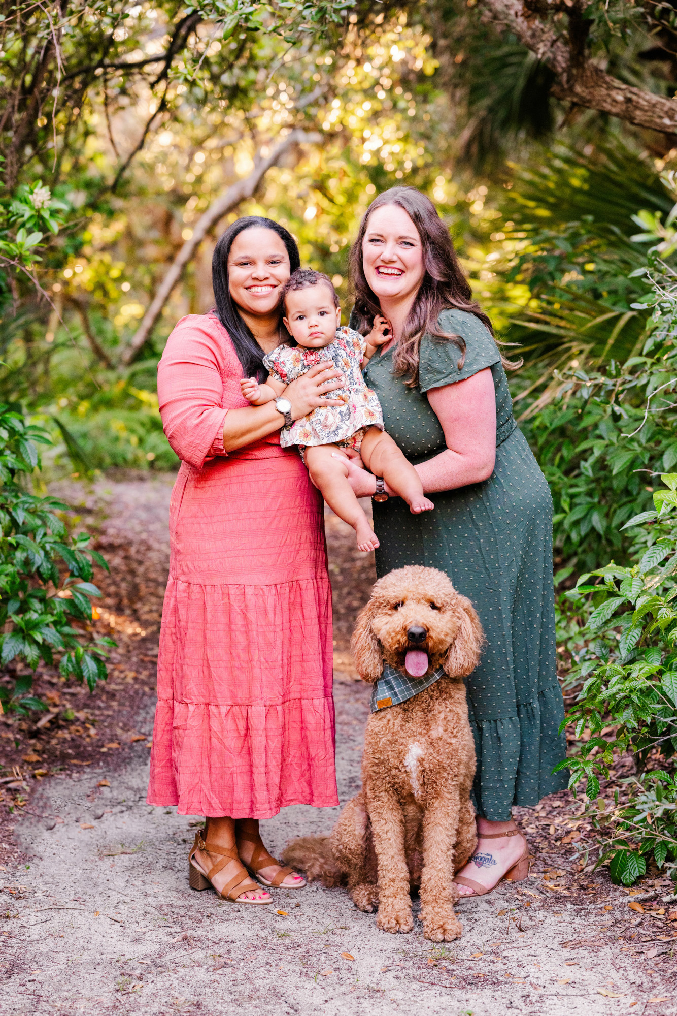 Two mothers smiling with their baby and curly-haired dog on a tropical garden path.